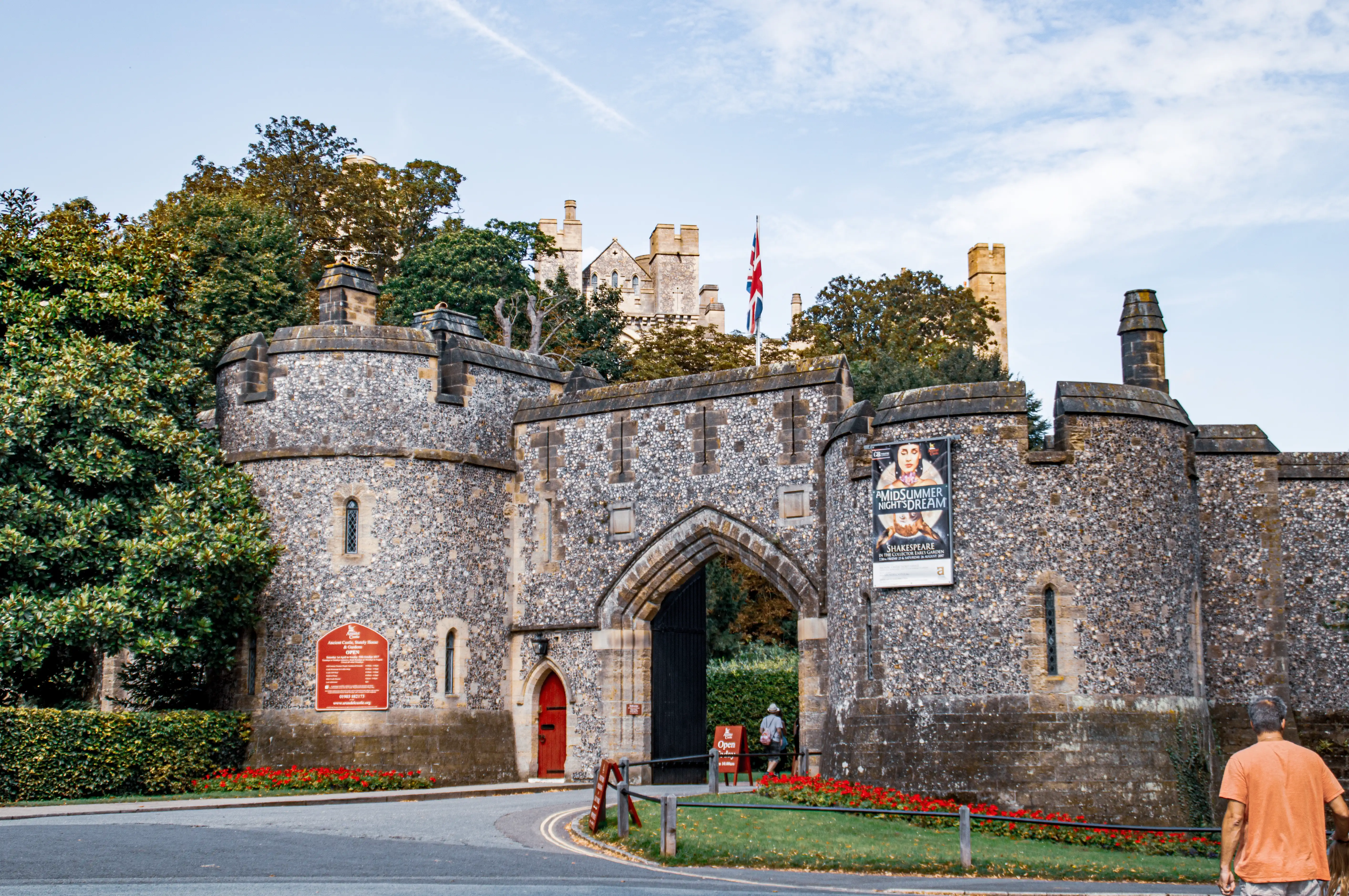 A medieval castle gateway made of stone and flint, with rounded towers featuring crenellated tops and a central archway. A Union Jack flag flies atop the gateway, and a poster for 'A Midsummer Night’s Dream' by Shakespeare is displayed on the right tower. A red door, signboard with opening times, and lush greenery with red flowers frame the scene