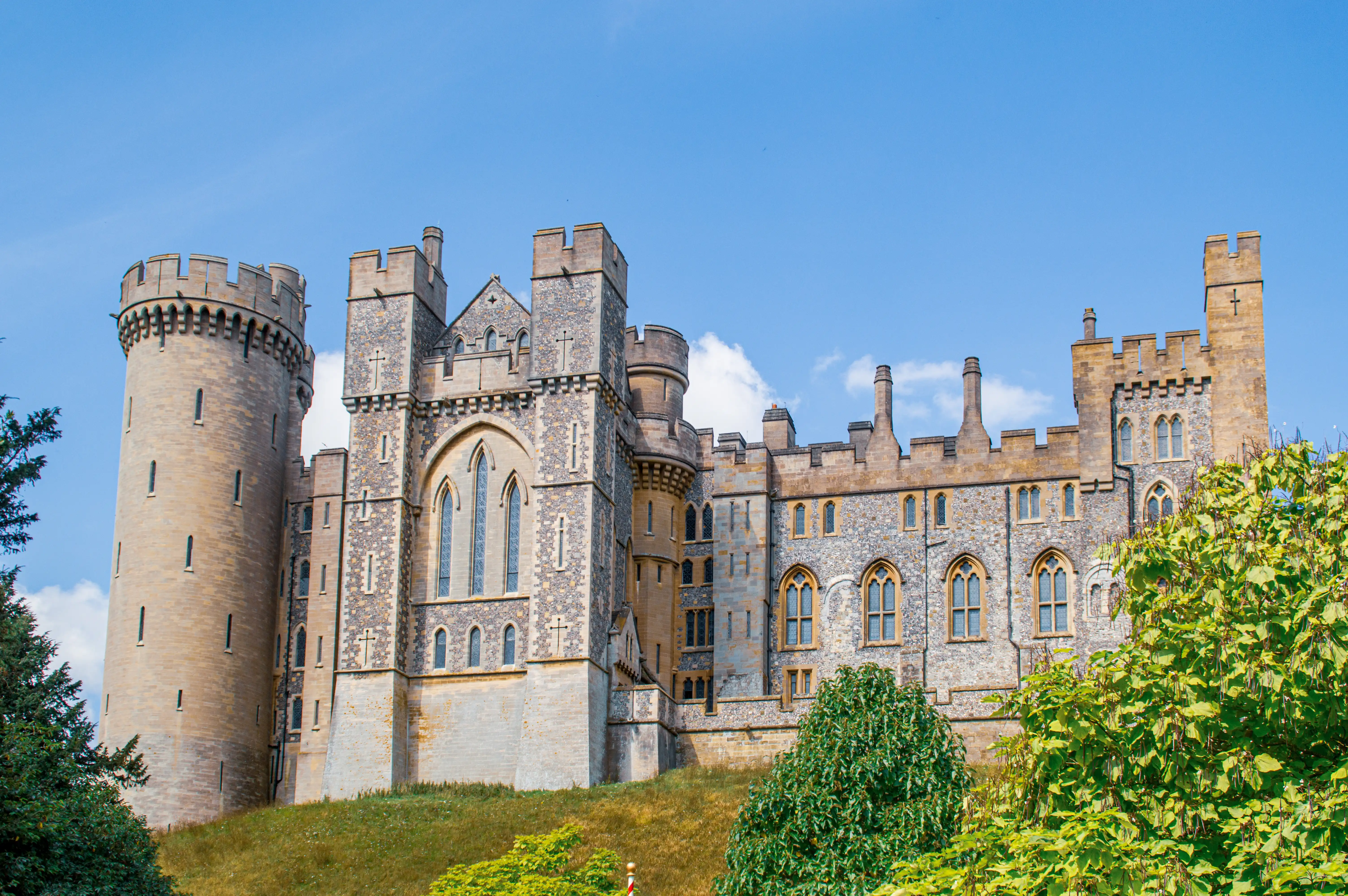 A wide shot of Arundel Castle in West Sussex, England, featuring its stone towers and battlements against a bright blue sky with some clouds. Green trees and grass surround the castle.