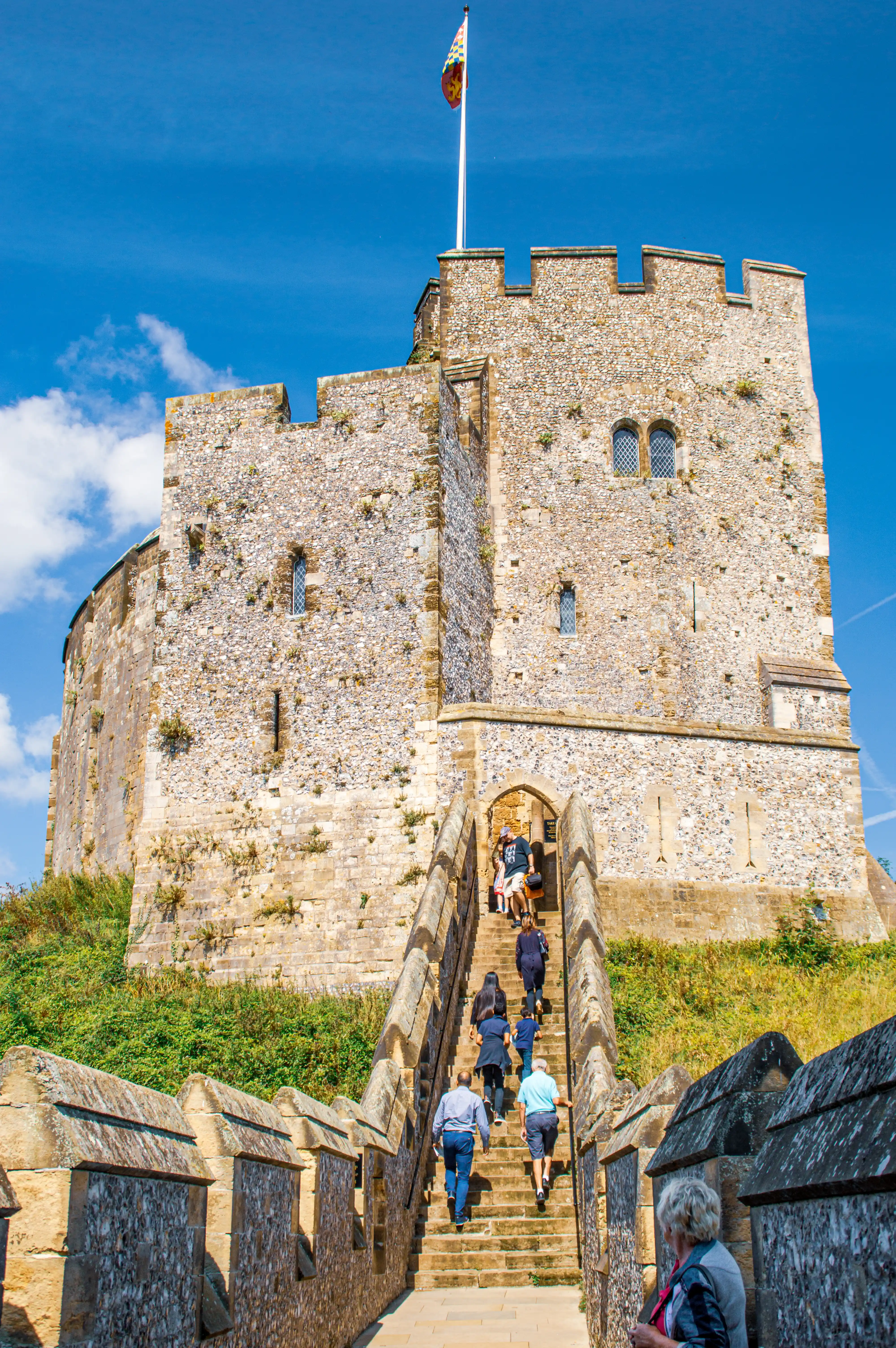 A party of people walks up a wide stone staircase towards the imposing stone keep of Dover Castle, which has a flag flying from its parapet.