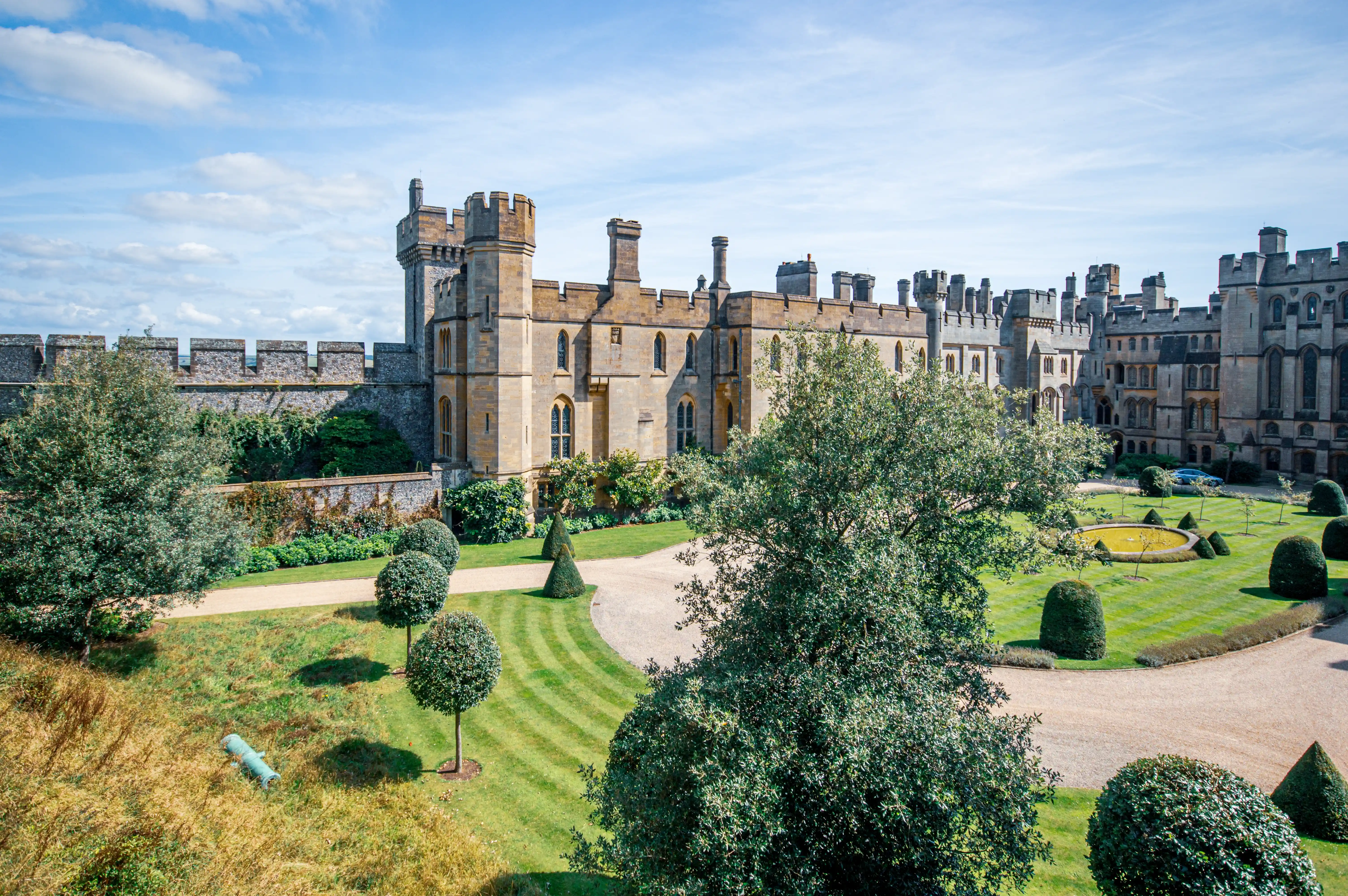 A wide-angle shot of a sprawling stone castle, featuring crenellated battlements and towers. In the foreground, immaculately kept lawns with striped patterns lead to a circular fountain. Various trees and shaped bushes are dotted across the grounds. A grey gravel driveway curves towards the castle.