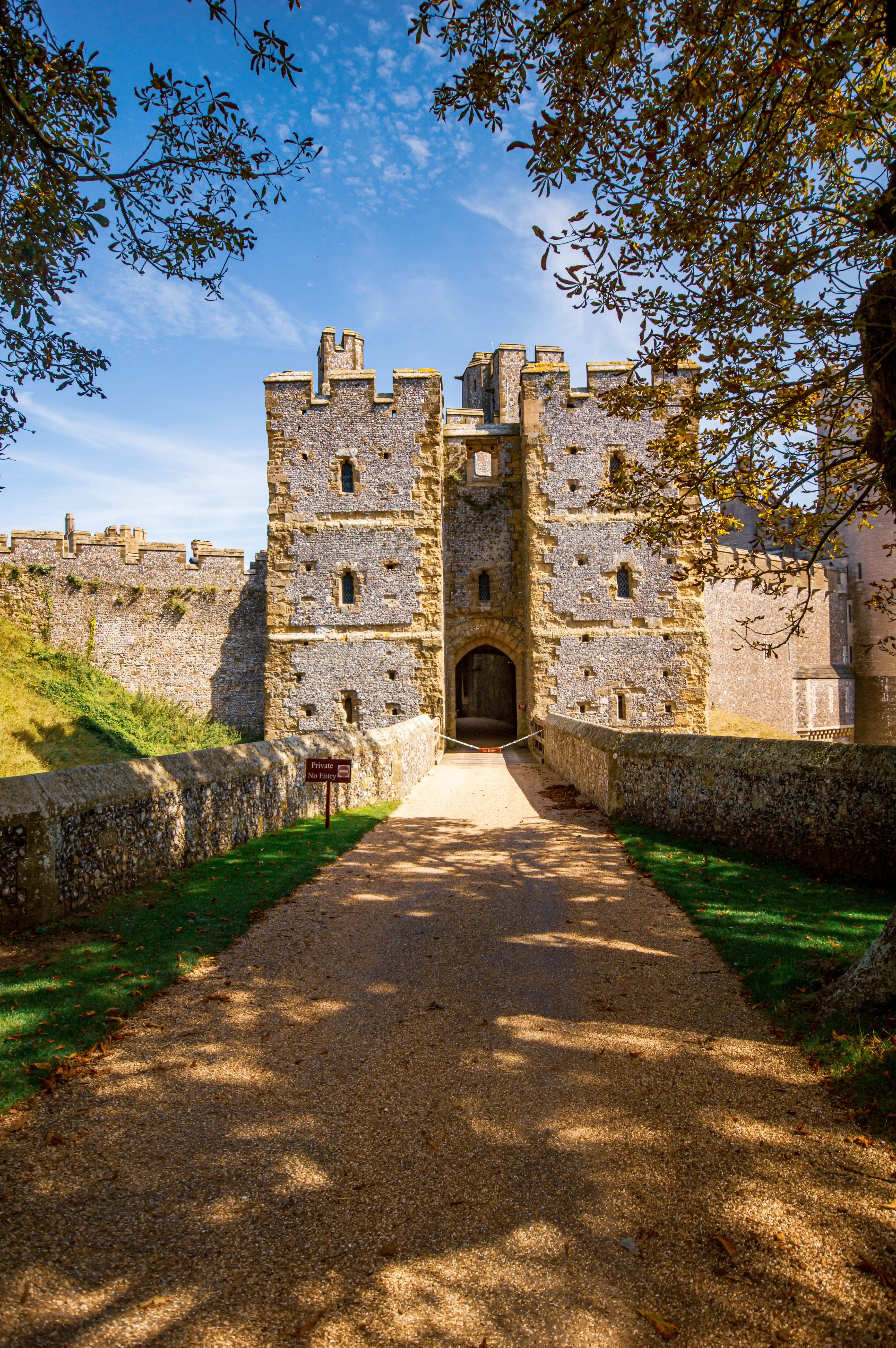 A gravel pathway approaches the fortified entrance of a medieval castle with stone walls and turrets, viewed under a bright blue sky and dappled tree shadows.