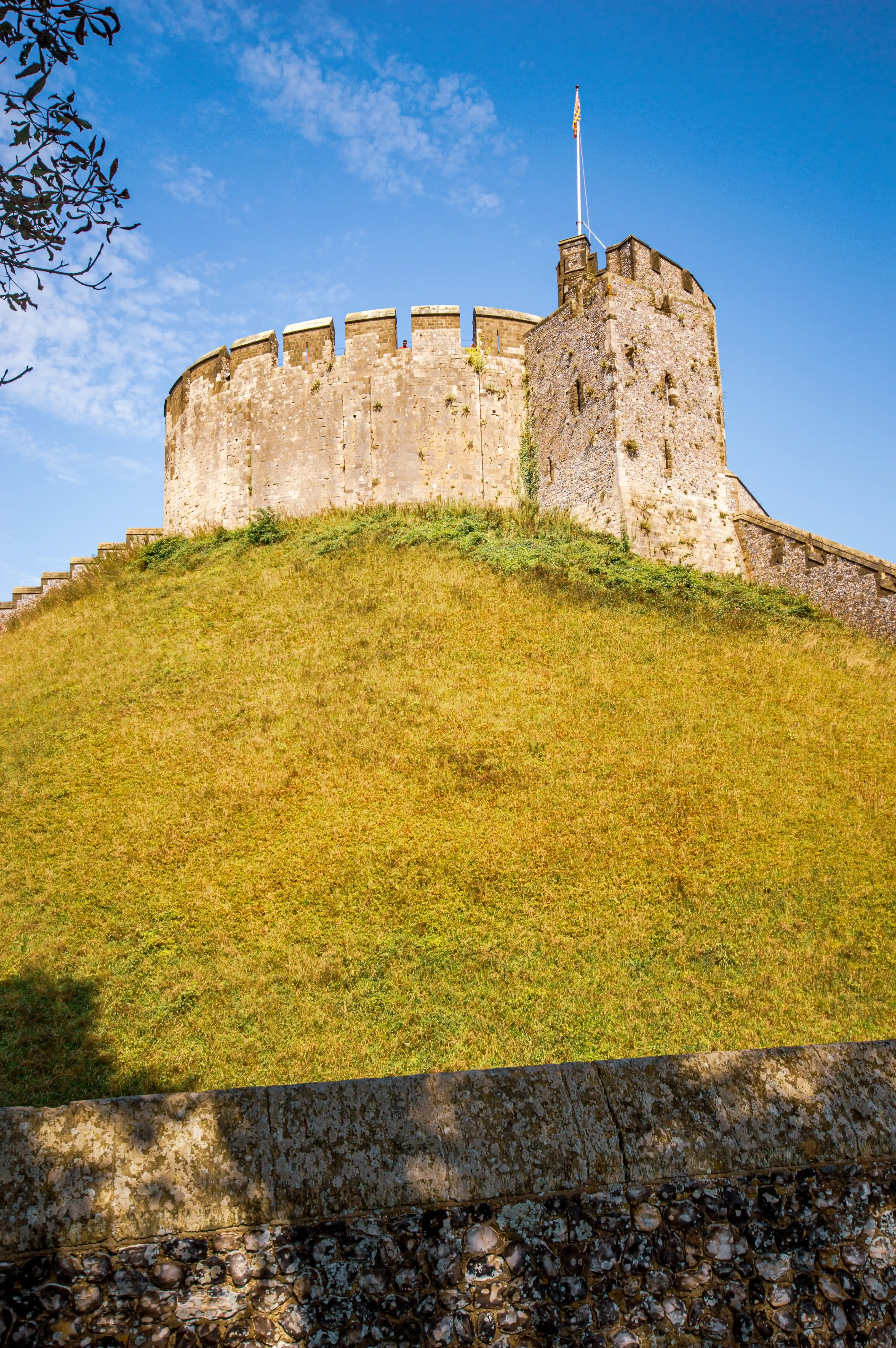 Medieval stone keep with battlements and a tall tower on a grassy mound under a clear blue sky.
