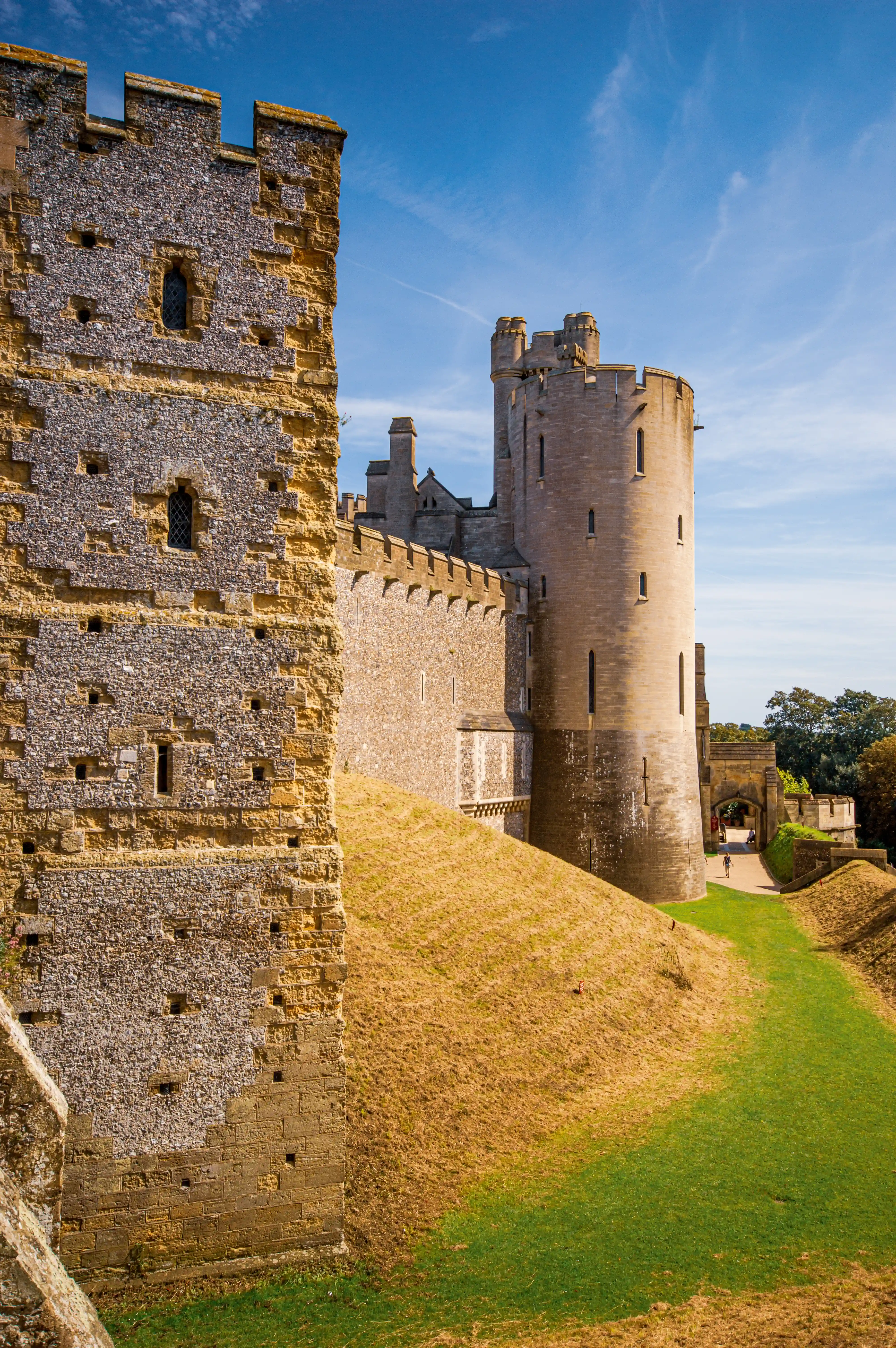 A stone castle tower with battlements stands prominently, viewed from the side of another stone wall. A grassy, sloped moat area with patches of dried grass leads towards an archway entrance. The sky is blue with scattered white clouds.