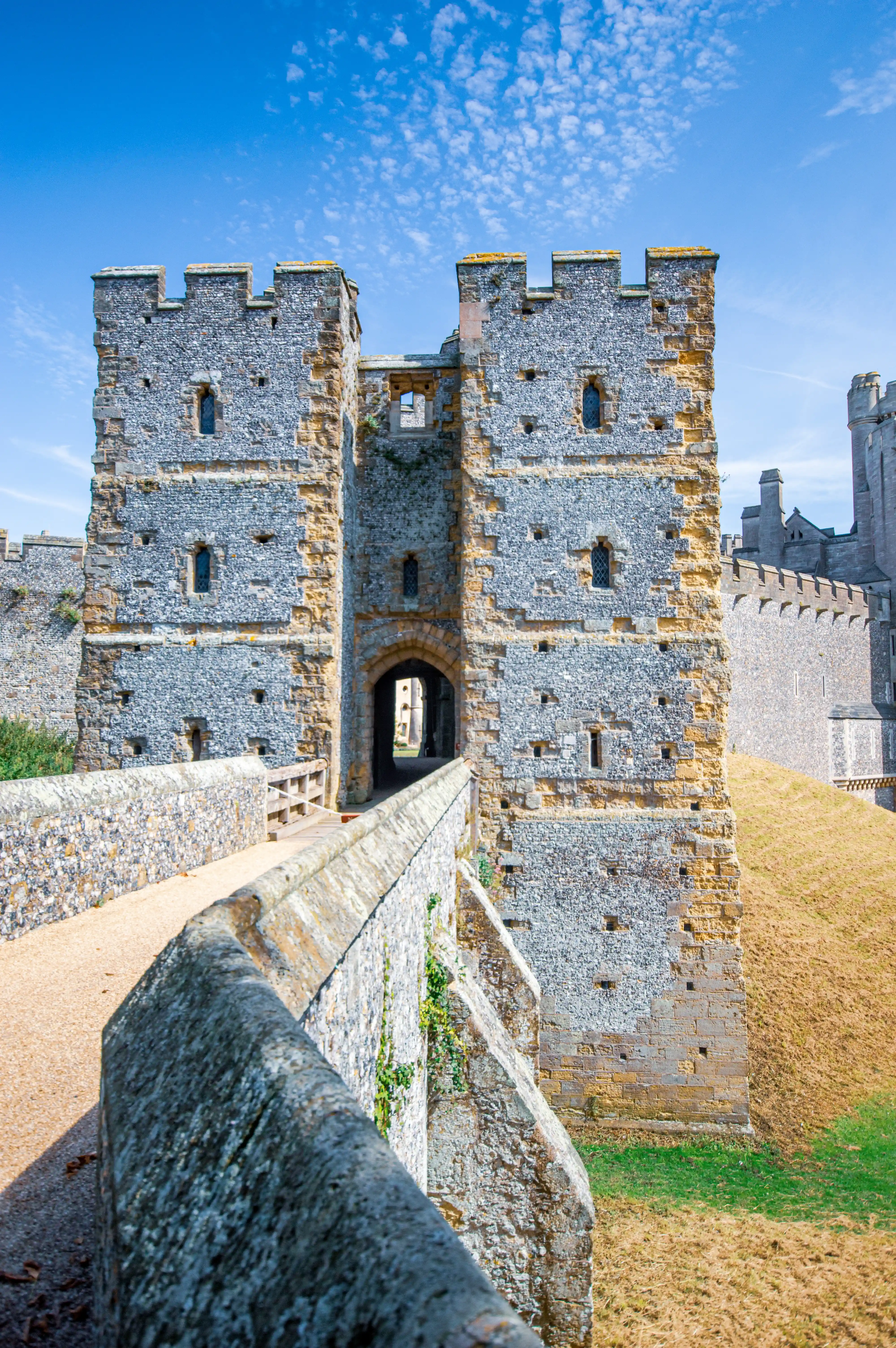 A stone bridge curves towards the imposing gateway of a historic castle, with crenellated towers and arched entrance. The scene is bathed in sunlight under a bright blue, partly cloudy sky.