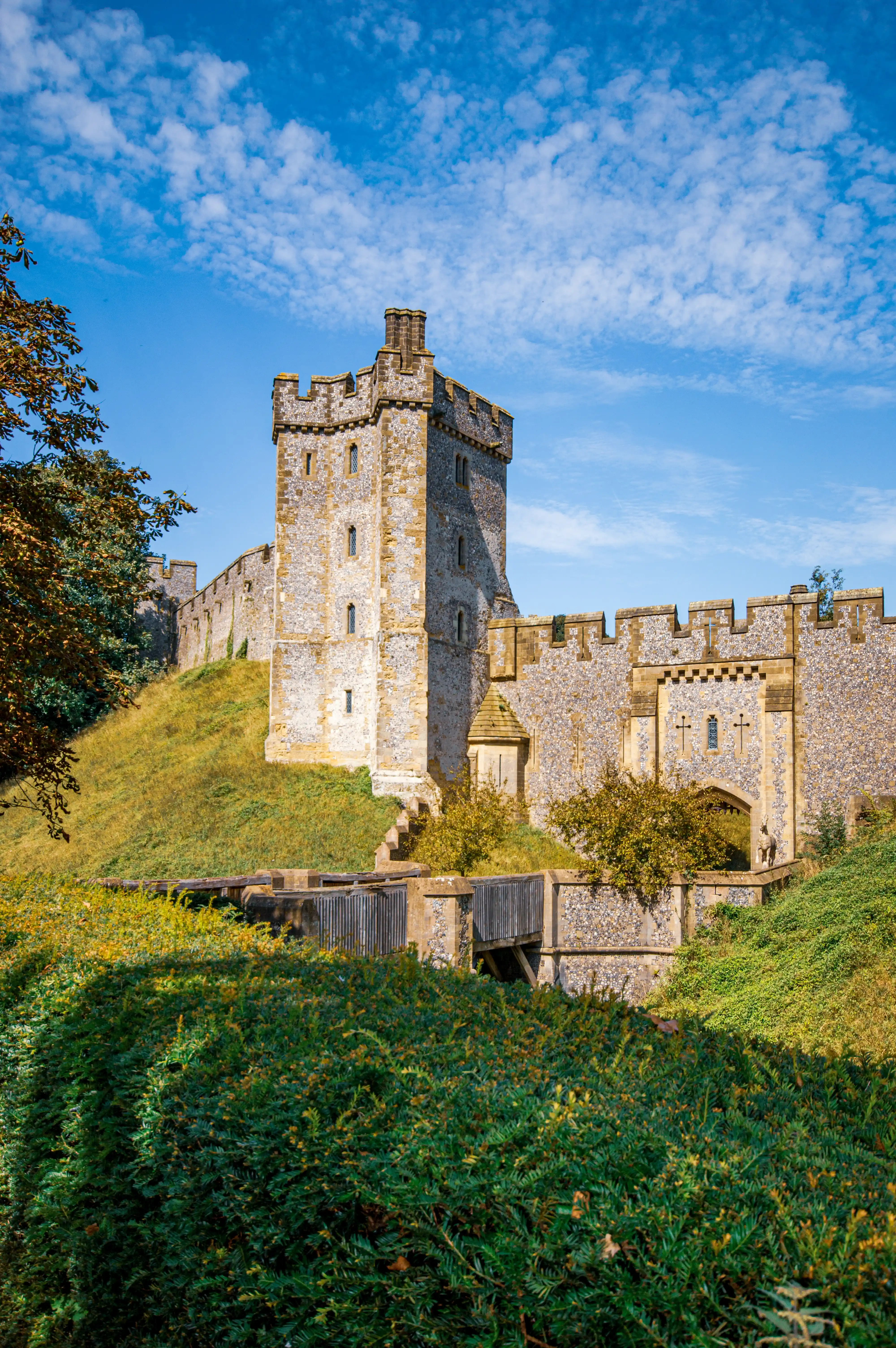 A medieval stone castle tower and walls on a grassy hill, with a wooden walkway in the foreground and a bright blue sky with clouds above.