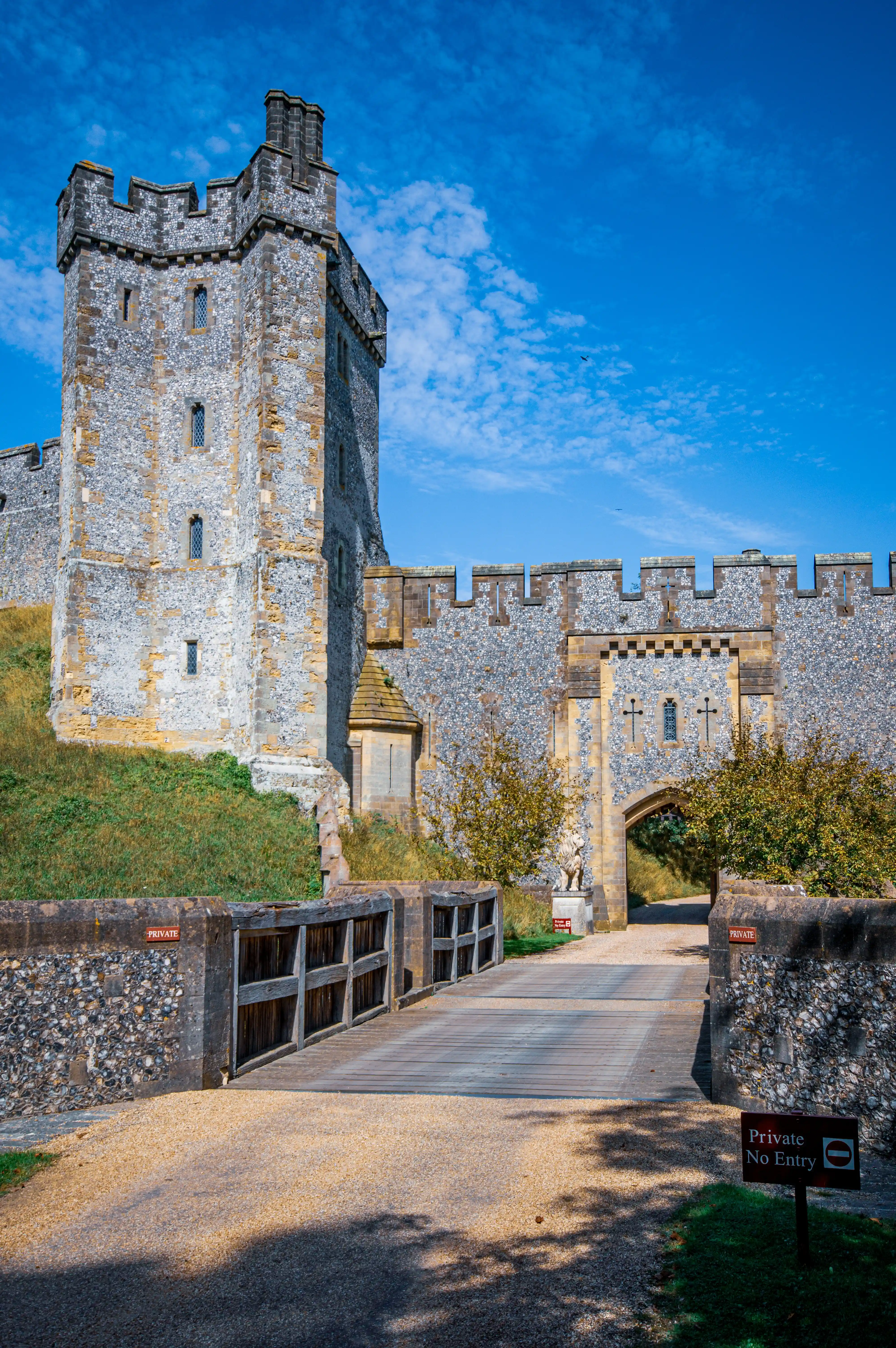 A stone castle drawbridge leads to an arched gateway of a historic castle. The castle walls and a tower are made of grey stone with some yellow stone accents. The sky is a vibrant blue with scattered white clouds.