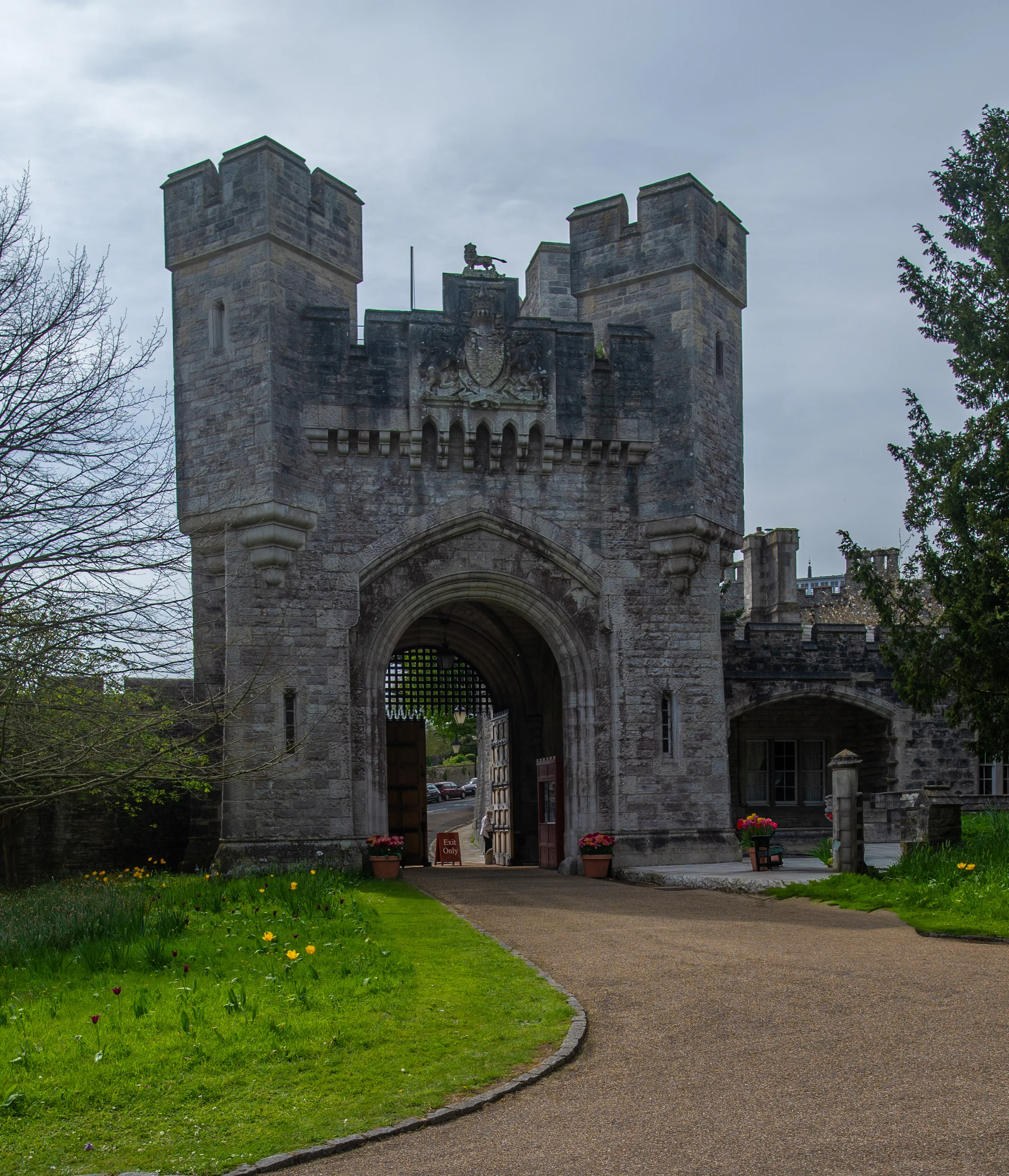 two towers featuring crenellated tops and a central archway secured by a metal portcullis. A coat of arms is displayed above the archway, and the gateway is surrounded by greenery, including grass, trees, and potted flowers.