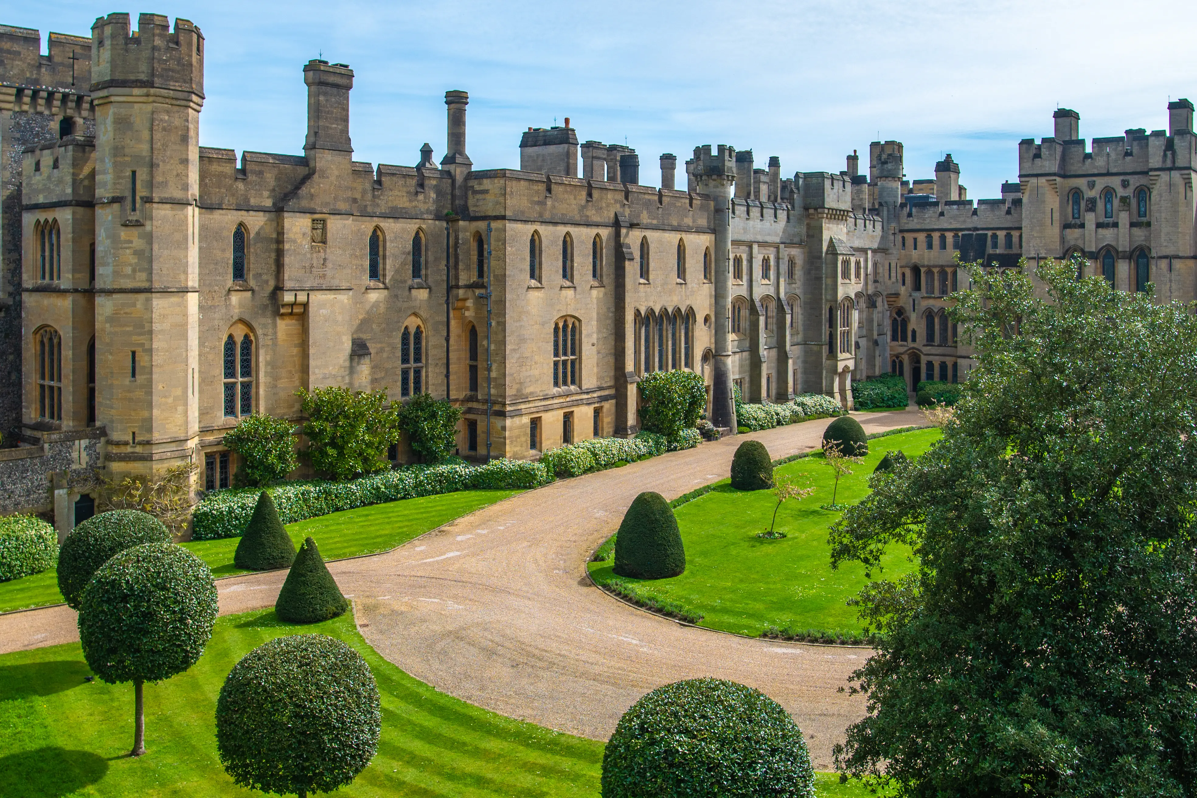  A wide view of a large, historic stone castle with turrets and many windows, seen from a slightly elevated position. In front of the castle is a curving gravel driveway surrounded by manicured lawns and perfectly shaped bushes, including spheres and cones. The sky is a pale blue with some cloud.