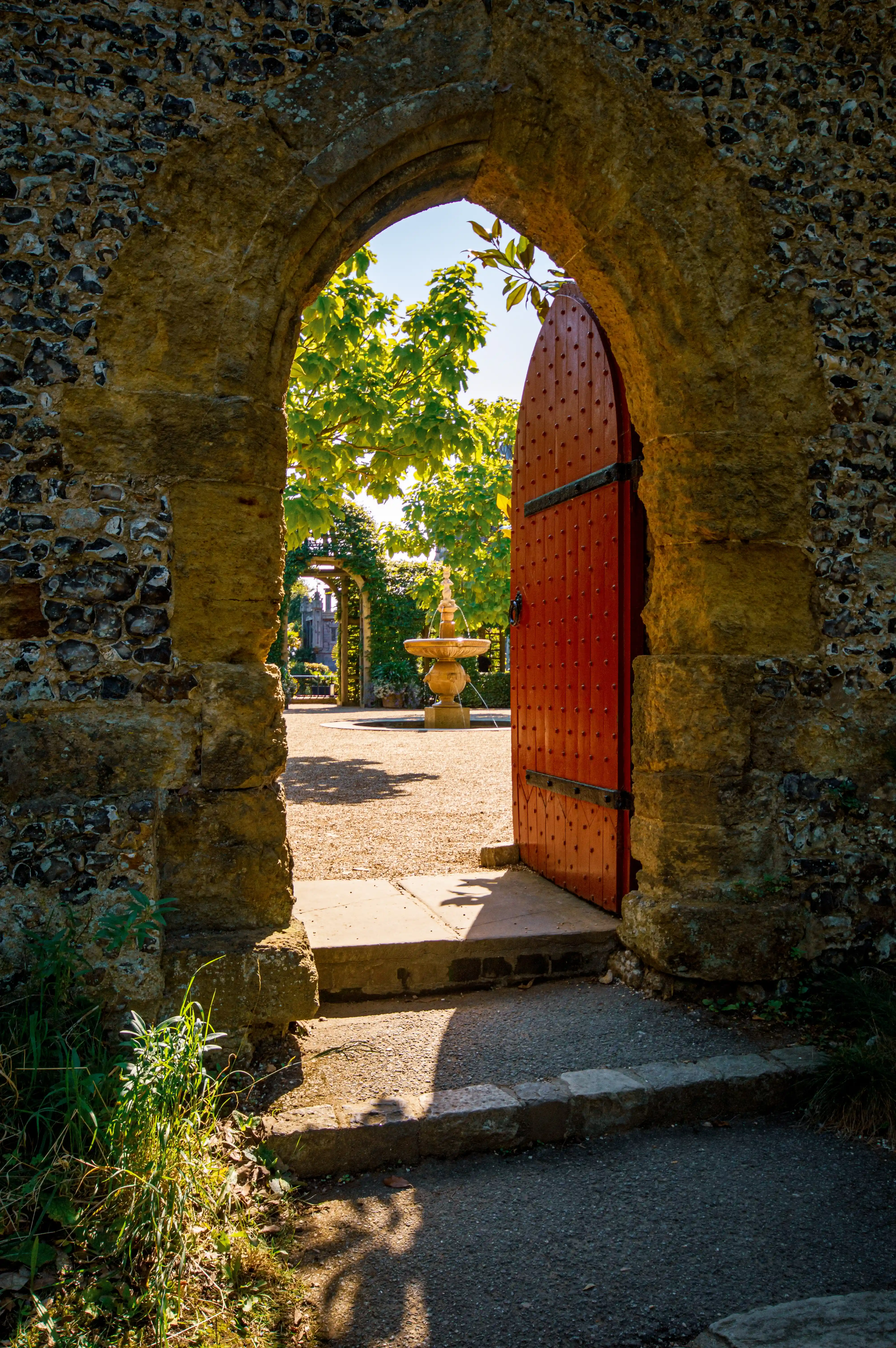 A stone archway with a pointed arch frames an open red wooden door. Beyond the door, a sunlit garden with a fountain is visible. Steps lead up to the doorway.