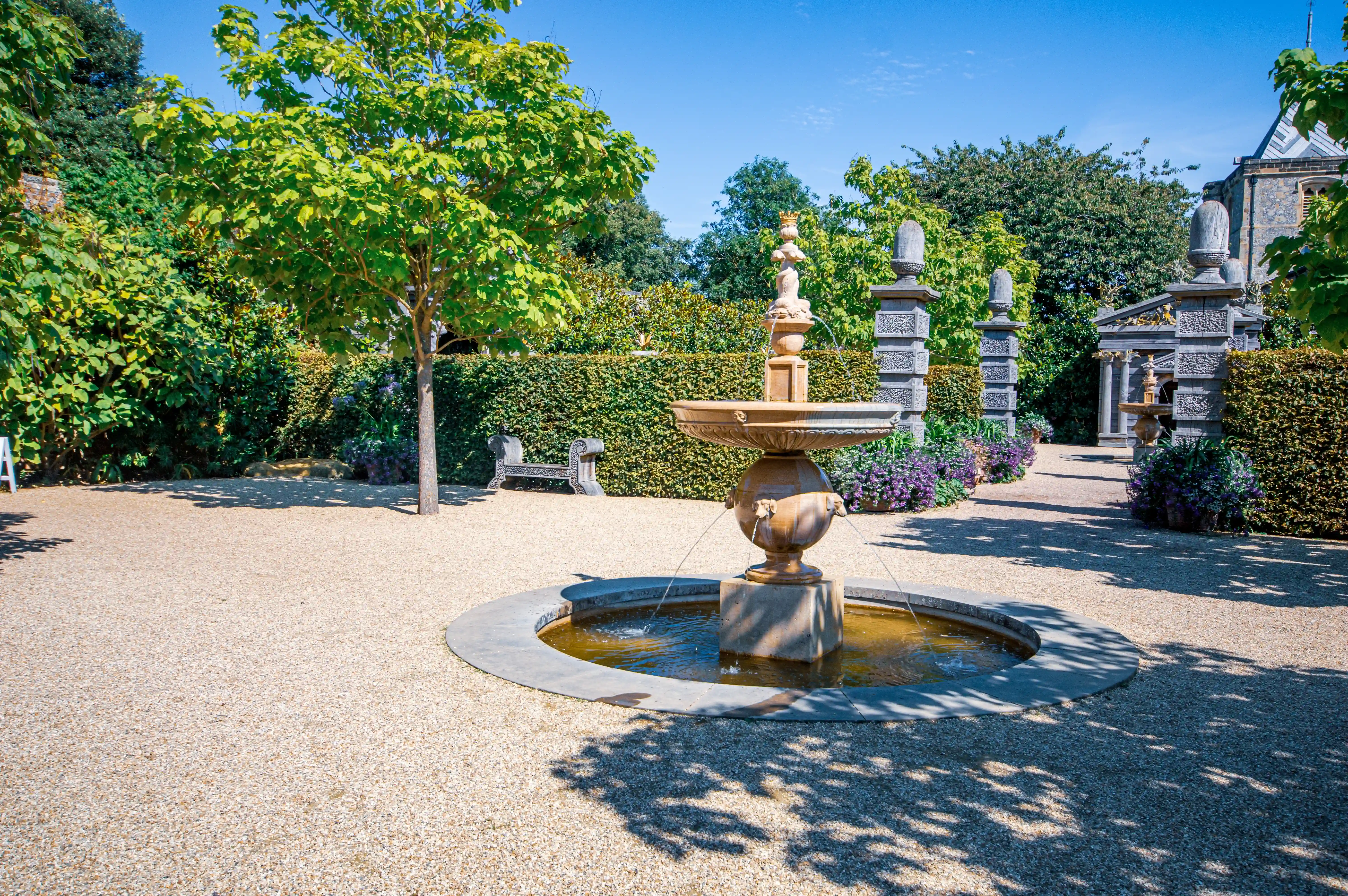 A stone fountain with water cascading down its levels, situated in a gravelled garden area. The background includes manicured hedges, a young tree, and architectural elements of a building.