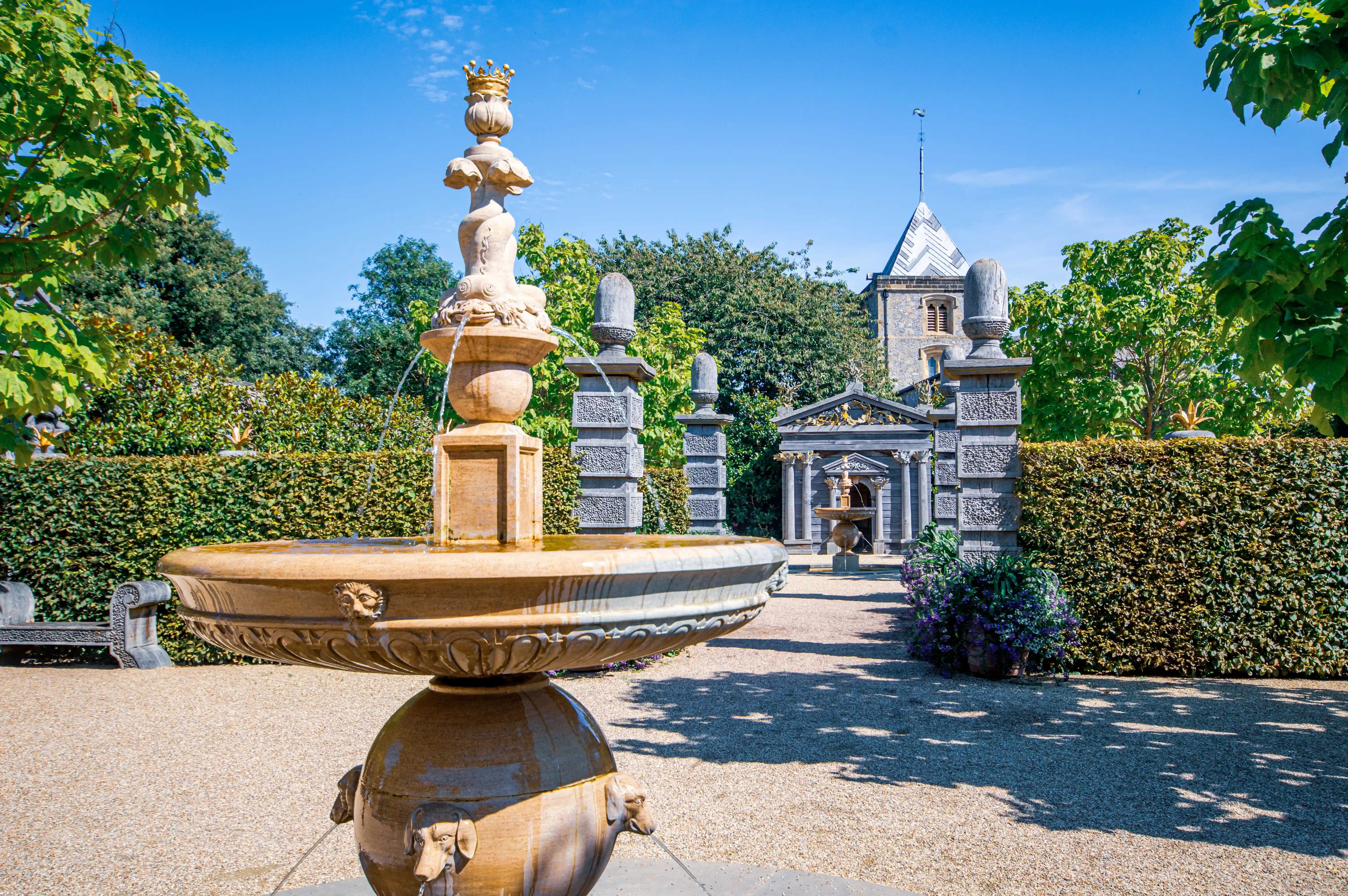 A stone fountain with flowing water in a formal garden, with a classical folly and a church tower in the background under a clear blue sky.