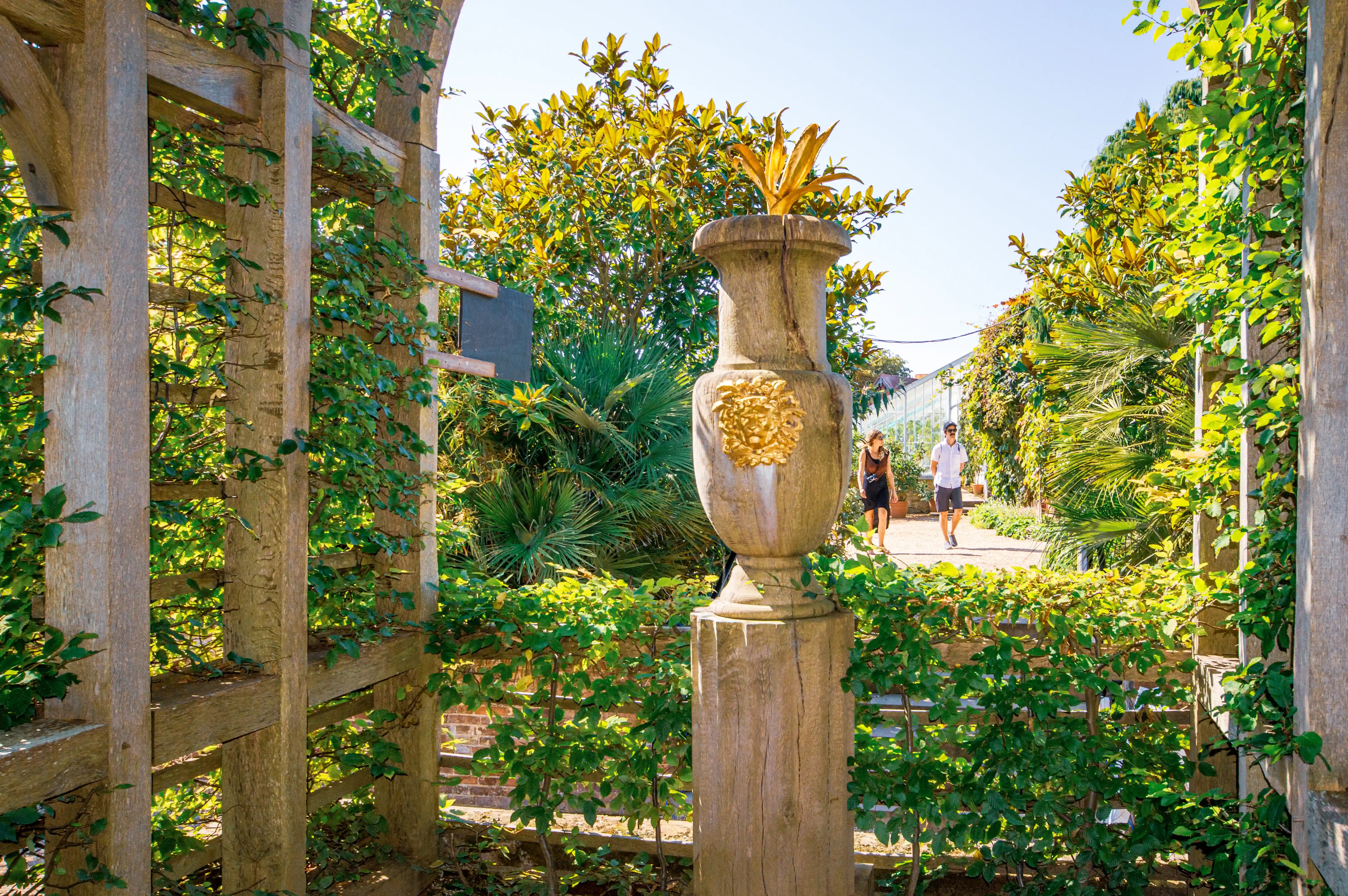 A stone urn with a gold emblem is in the foreground of a garden path. A couple walks away from the camera along the path, surrounded by abundant green foliage and trees. A wooden trellis structure is visible on the left.
