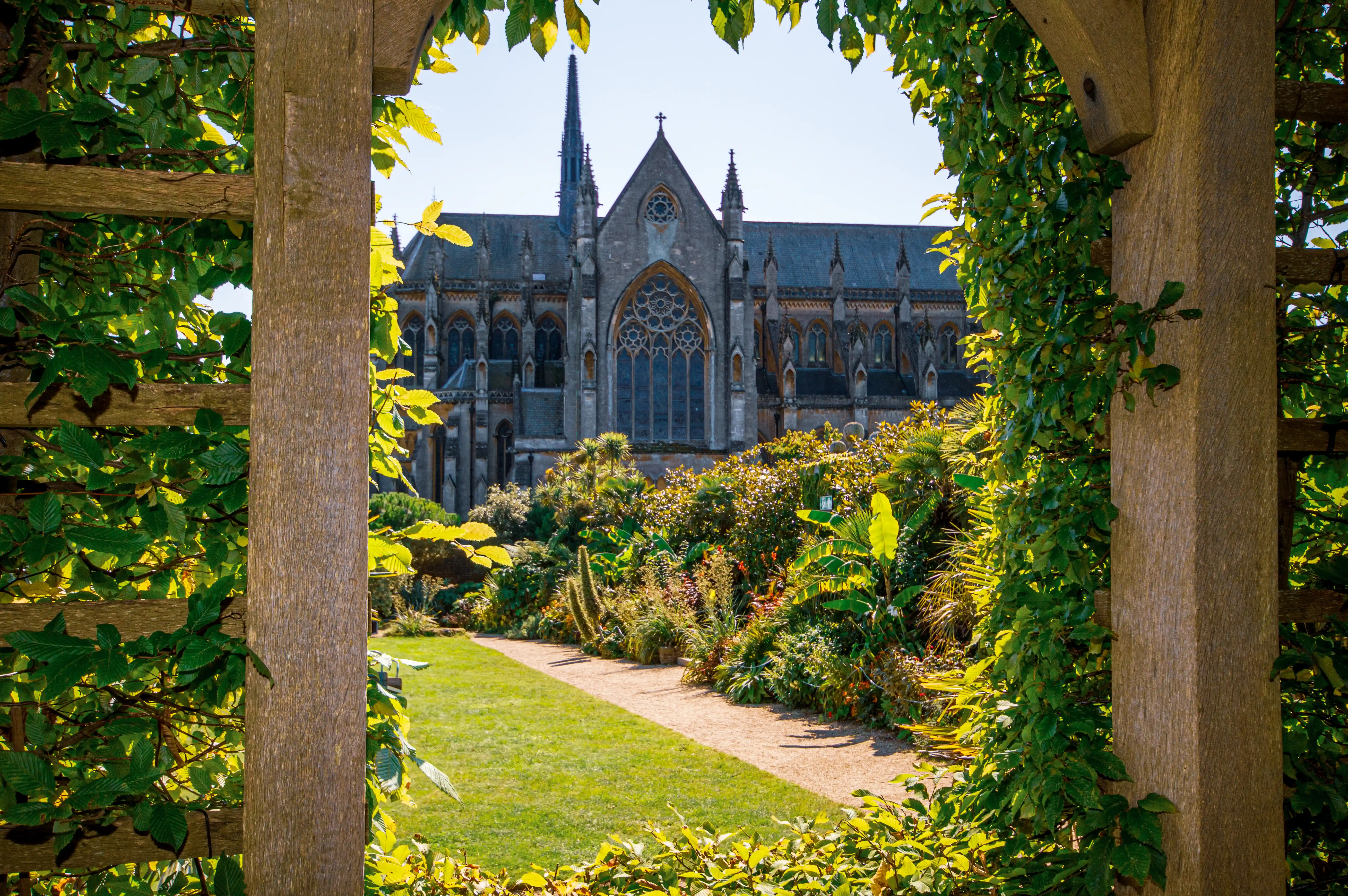 Through an archway draped in leaves, a large Gothic-style cathedral with a tall spire is visible, set against a clear blue sky. In front of the cathedral is a vibrant, densely planted garden with a gravel path leading through it.