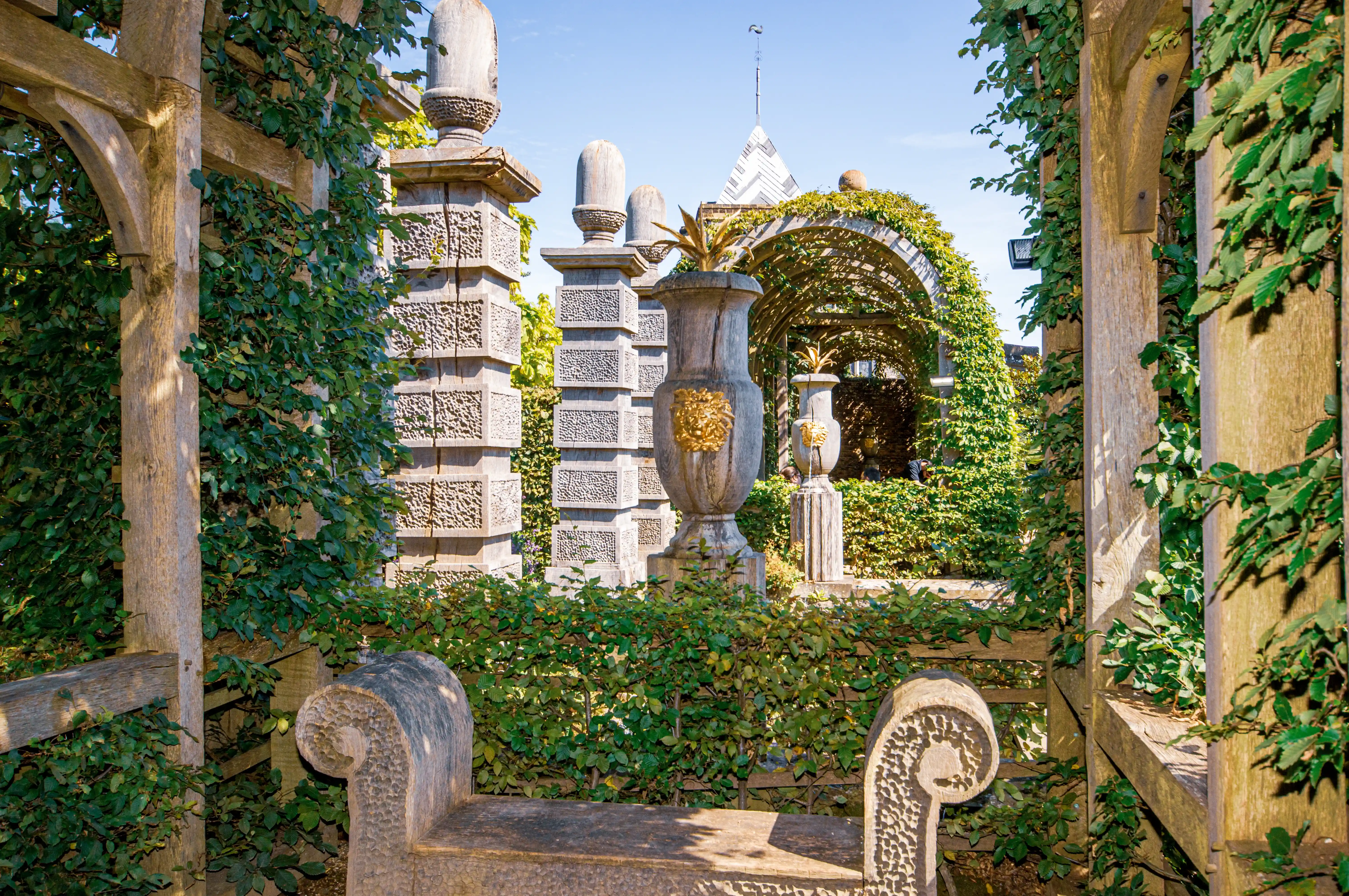 A stone bench sits in a verdant garden, framed by wooden trellises covered in ivy. In the mid-ground, carved stone pillars and a grand urn with a gold lion motif stand before an arched pergola heavily draped in green foliage. The scene is illuminated by bright sunlight under a blue sky.