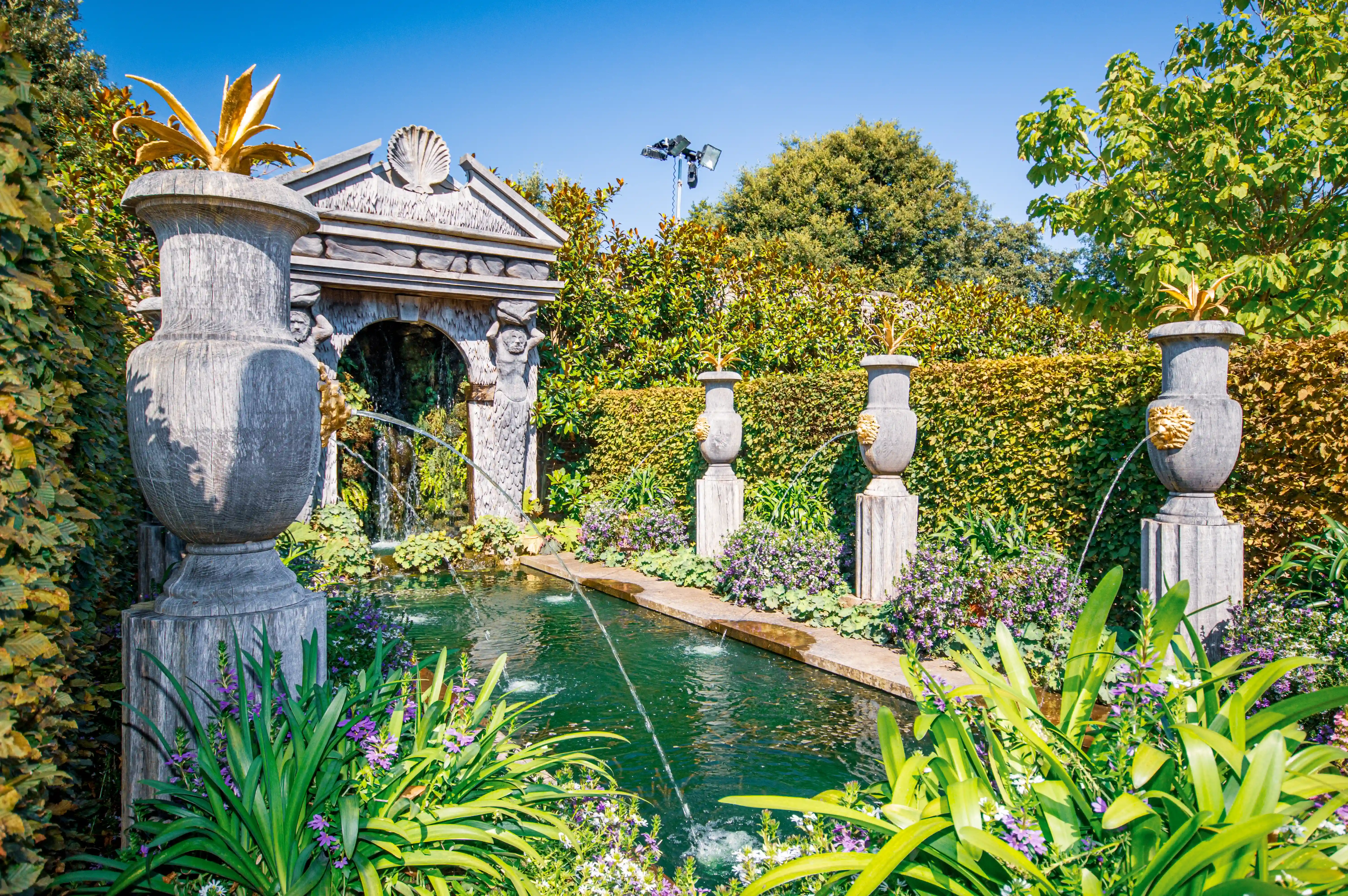A formal garden scene with a rectangular pond featuring several water jets. On the left, a large stone urn topped with a golden leaf-like ornament stands beside an ivy-covered wall. To the right of the pond, two more stone urns, each with a golden lion's head spout and a similar golden top ornament, stand on plinths. Behind the pond is an arched stone structure resembling a folly, with decorative carvings. The garden is enclosed by high, neatly trimmed hedges.