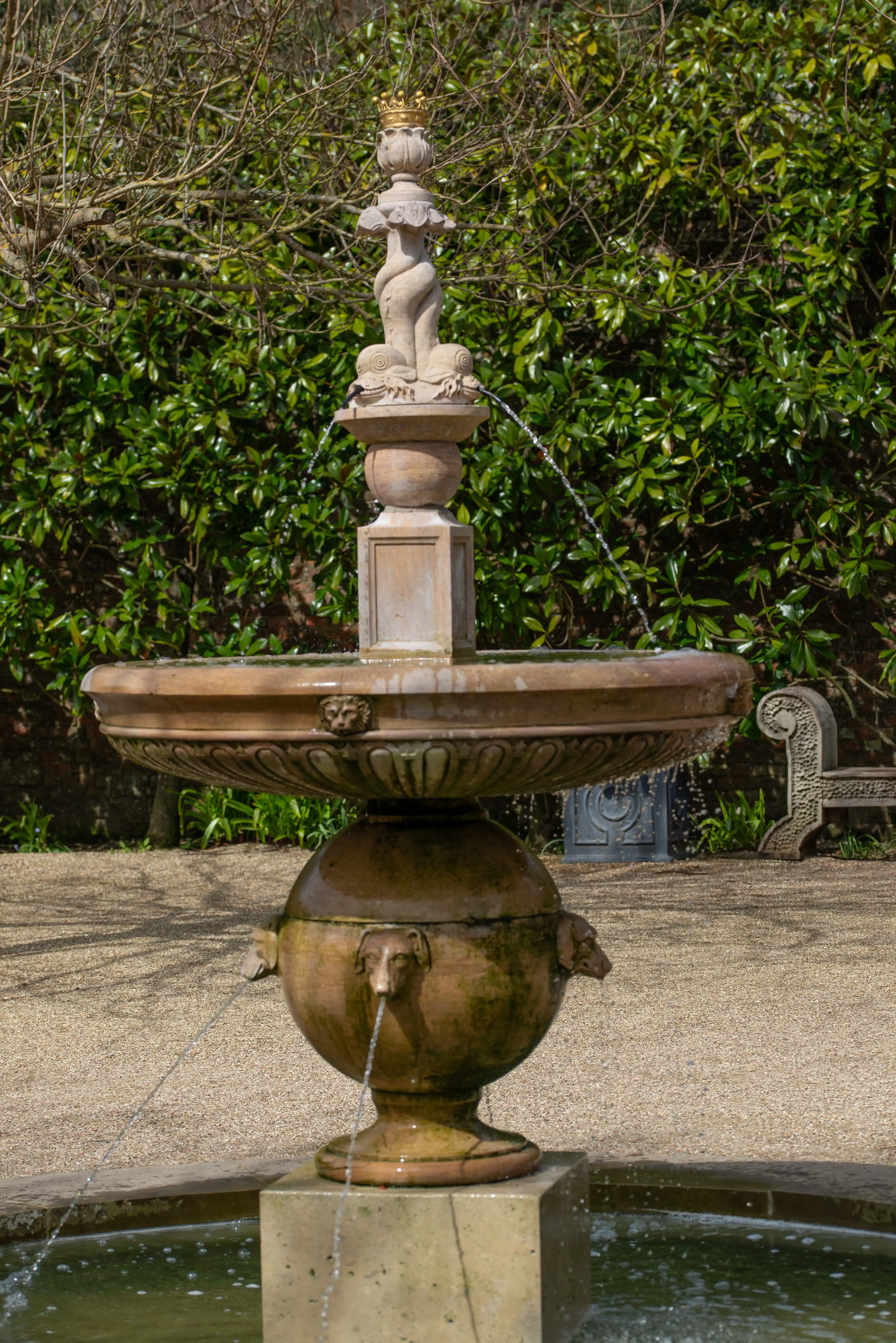 A tiered stone fountain with a central sculpture crowned with gold, spouting water from lion and dog-like heads, stands in a garden with a green hedge backdrop and a gravel path.