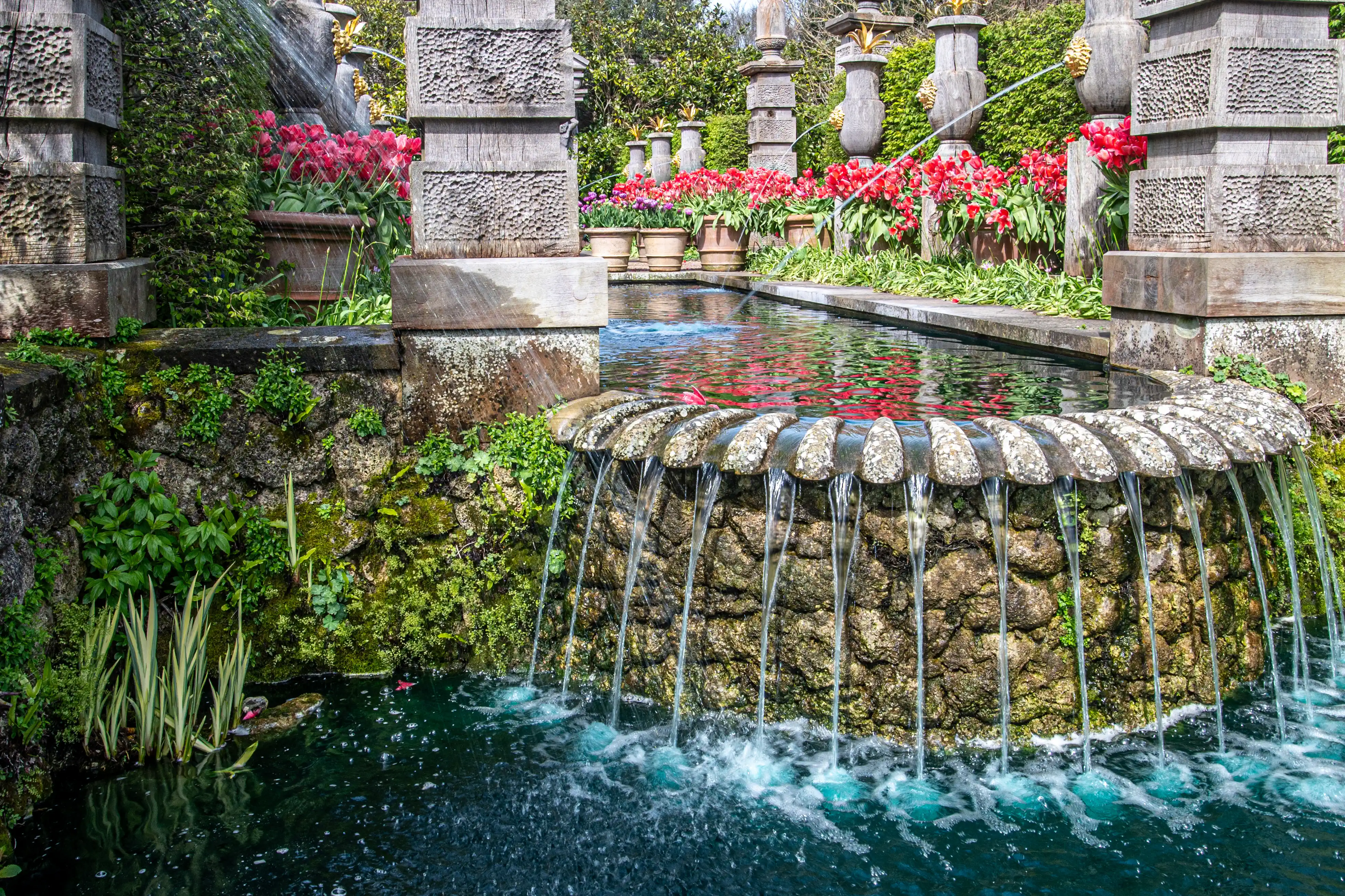 A stone fountain in a formal garden, with water flowing over a decorative edge into a pool. Red flowers and green foliage are visible in the background, alongside carved 