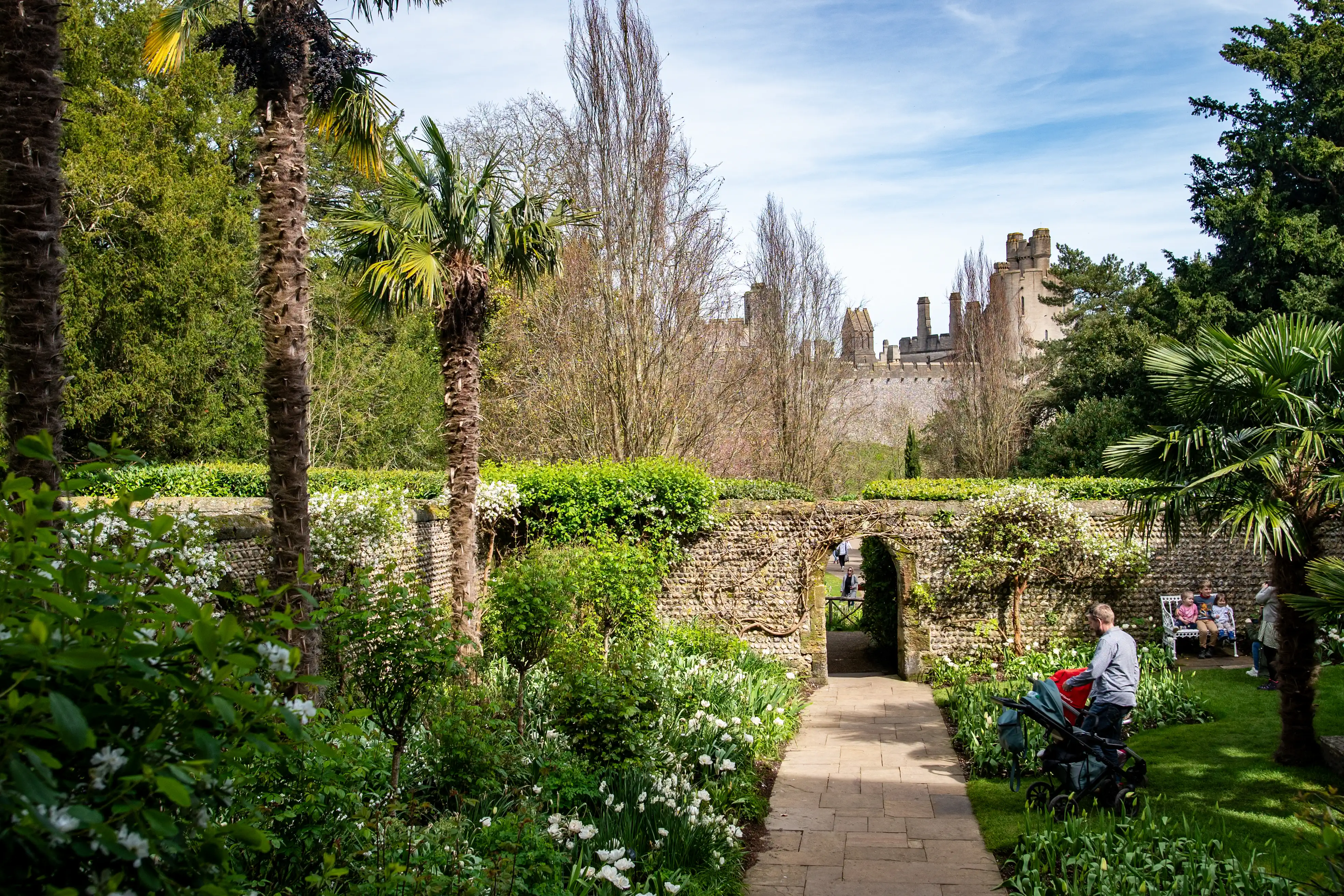 A stone pathway winds through a verdant garden with palm trees, leading to an arched stone opening. In the distance, a castle with crenellated towers stands against a partly cloudy blue sky.
