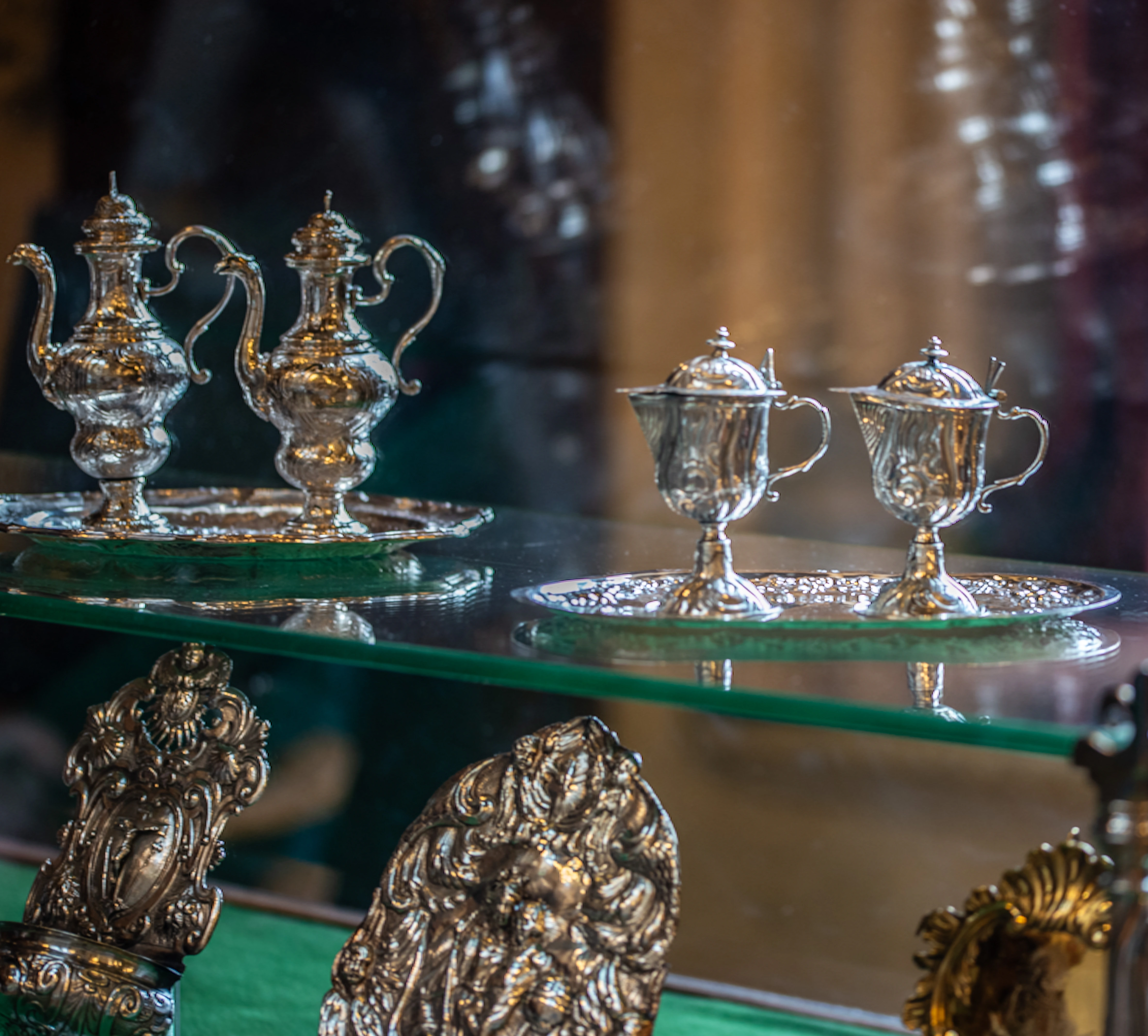 A close-up view of two pairs of vintage silver serving pieces, including teapots or coffee pots and smaller lidded jugs, displayed on a glass shelf.