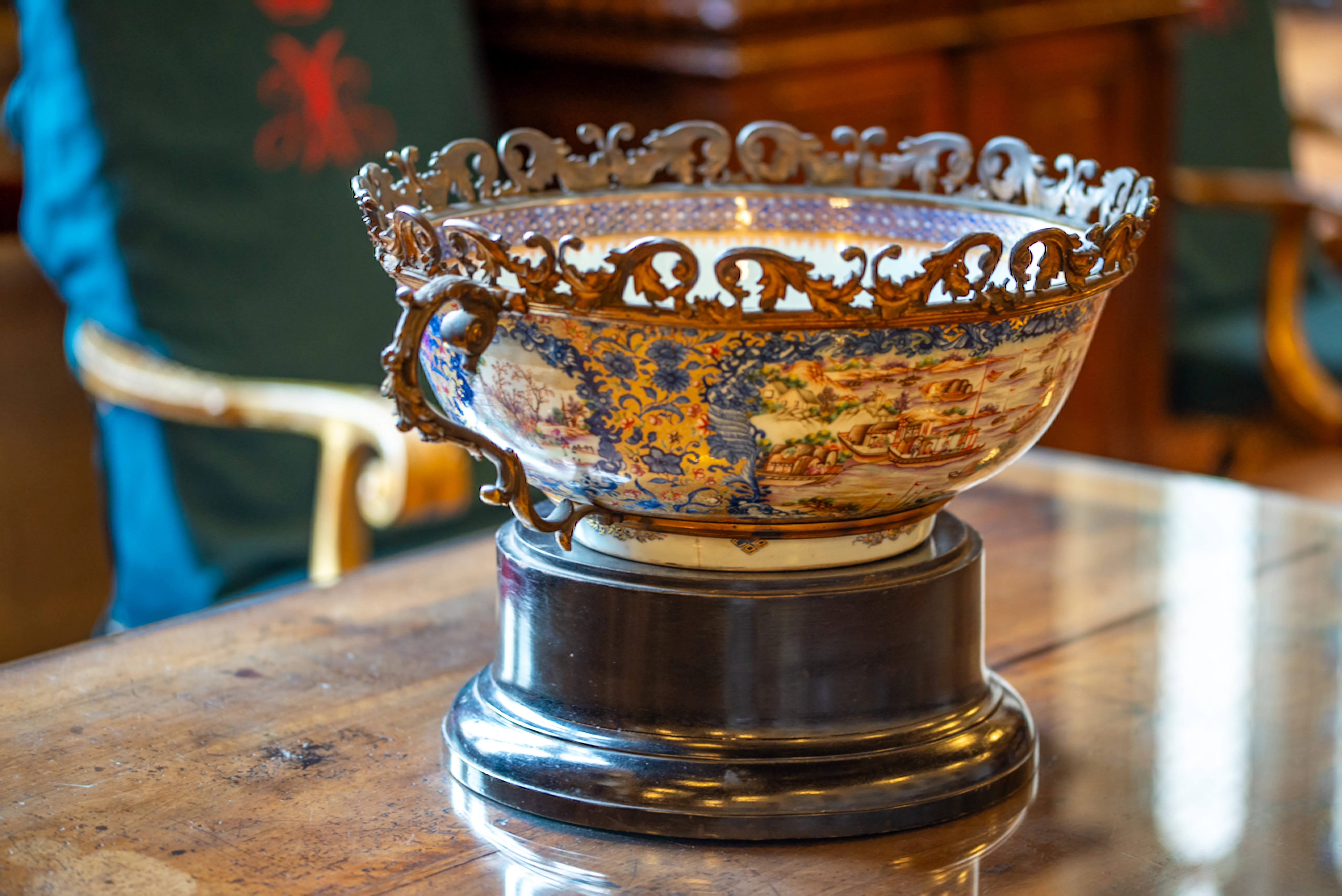 A close-up, eye-level shot of an ornate porcelain bowl with gilded metalwork, displaying detailed hand-painted scenes of boats and buildings. The bowl rests on a dark wooden pedestal on a polished wooden table.