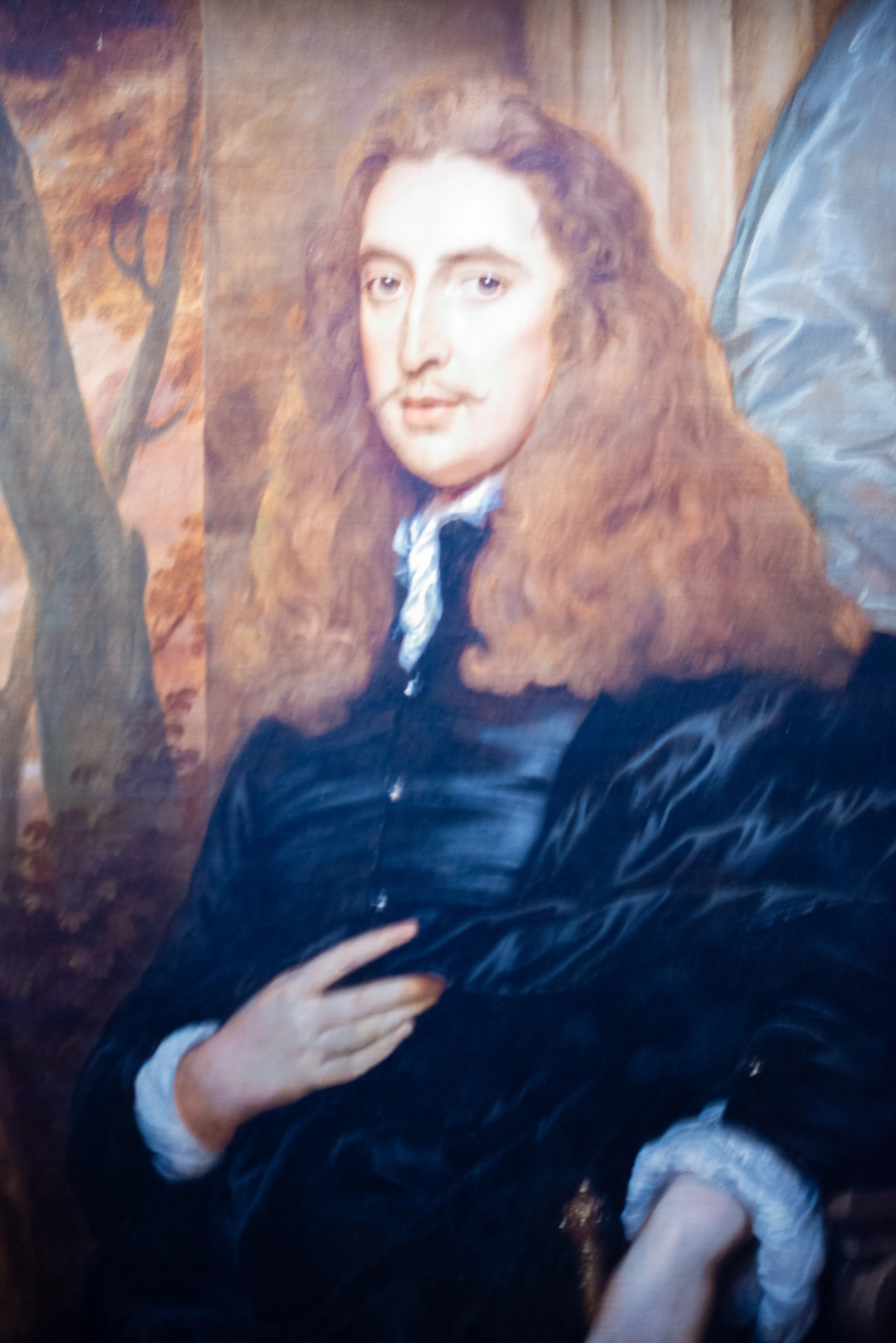 A painting of a man with long, wavy brown hair and a moustache, wearing a dark jacket and a white ruffled collar. He is shown in a half-length portrait against a backdrop of trees.