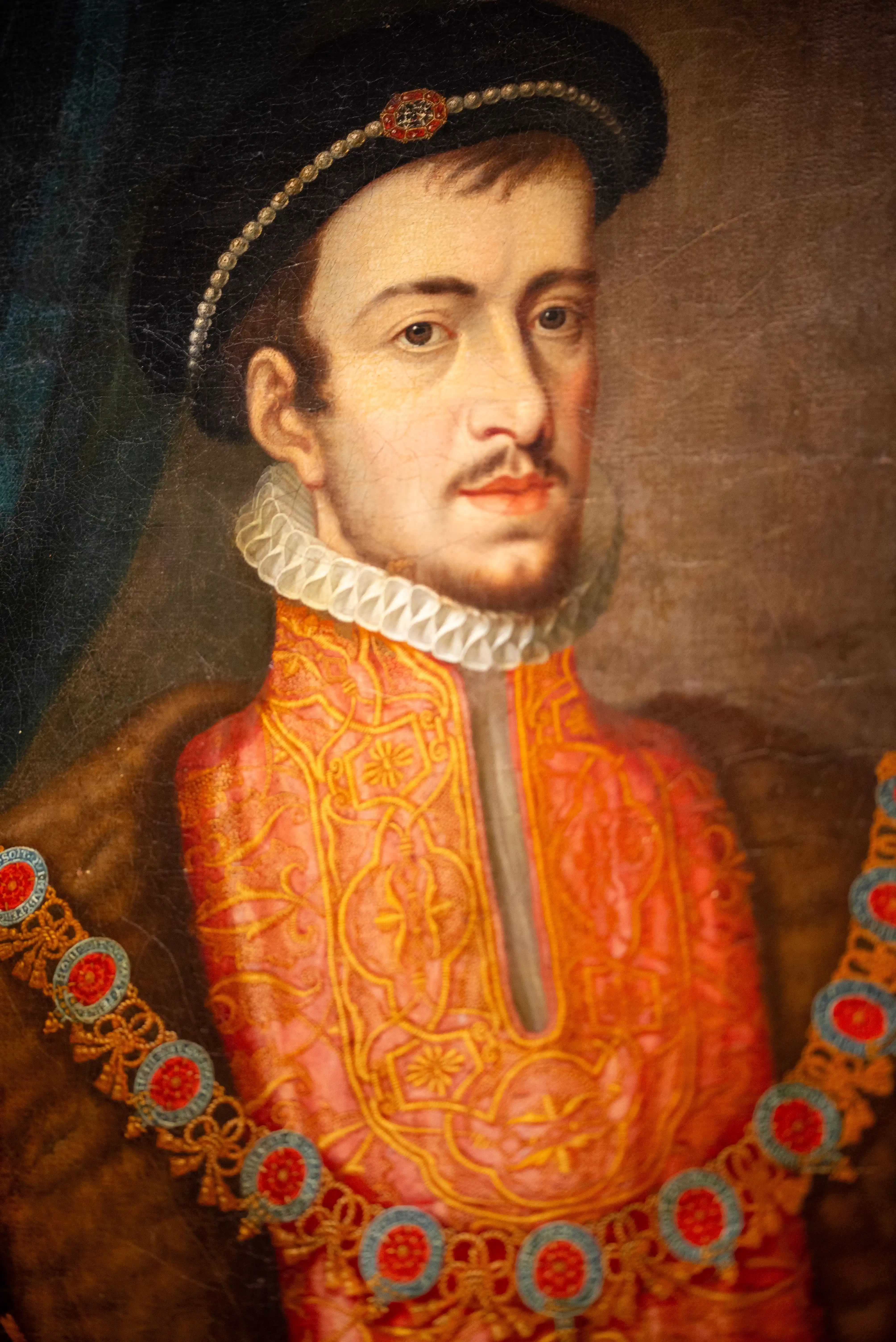 A close-up painting of a man in historical attire, featuring a black hat with a jewelled band, a white ruffled collar, and an ornate orange garment.