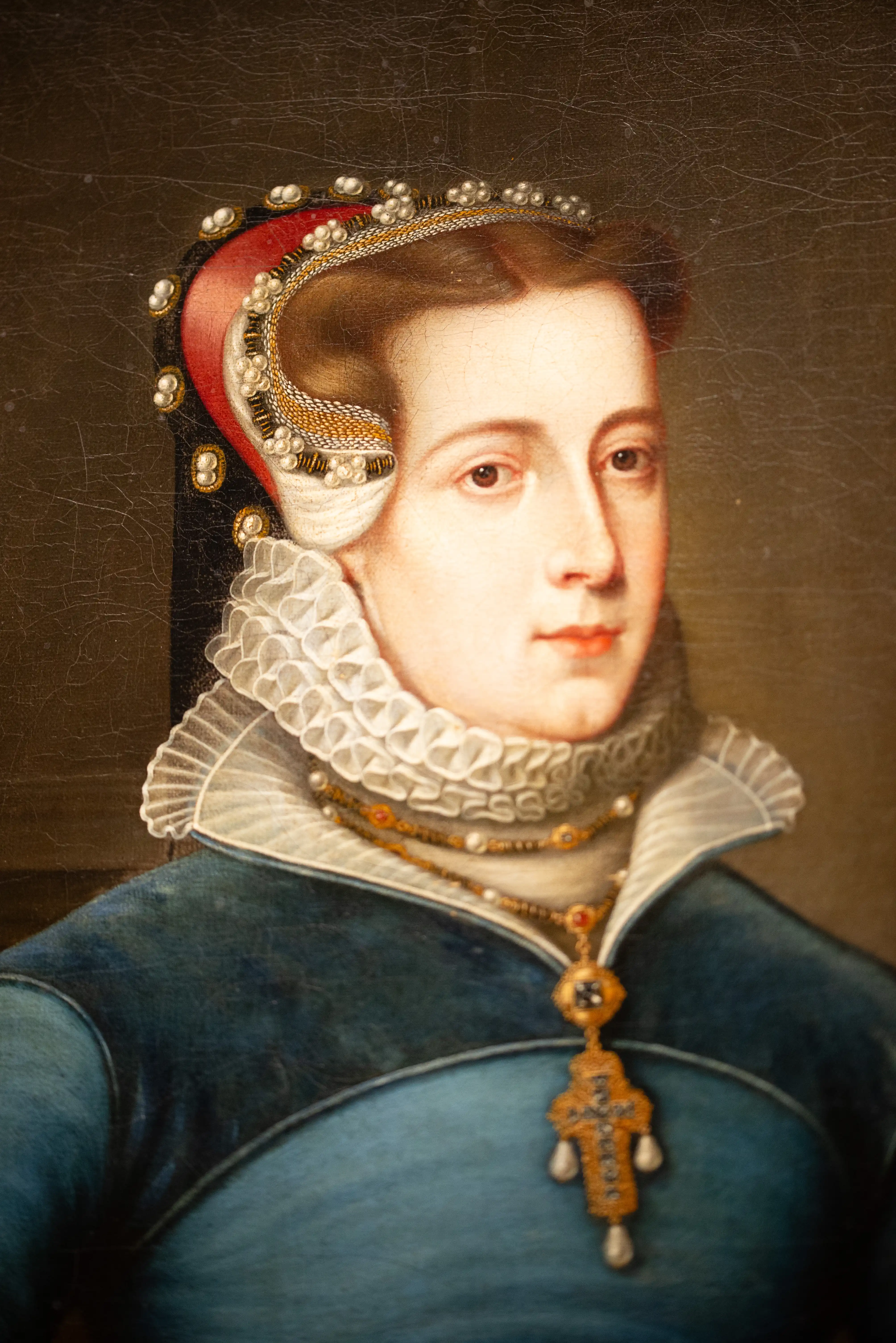 Close-up portrait painting of a woman wearing a jewelled headdress, a ruffled white collar, and a gold pendant necklace with pearls and gemstones. She is dressed in a blue garment.