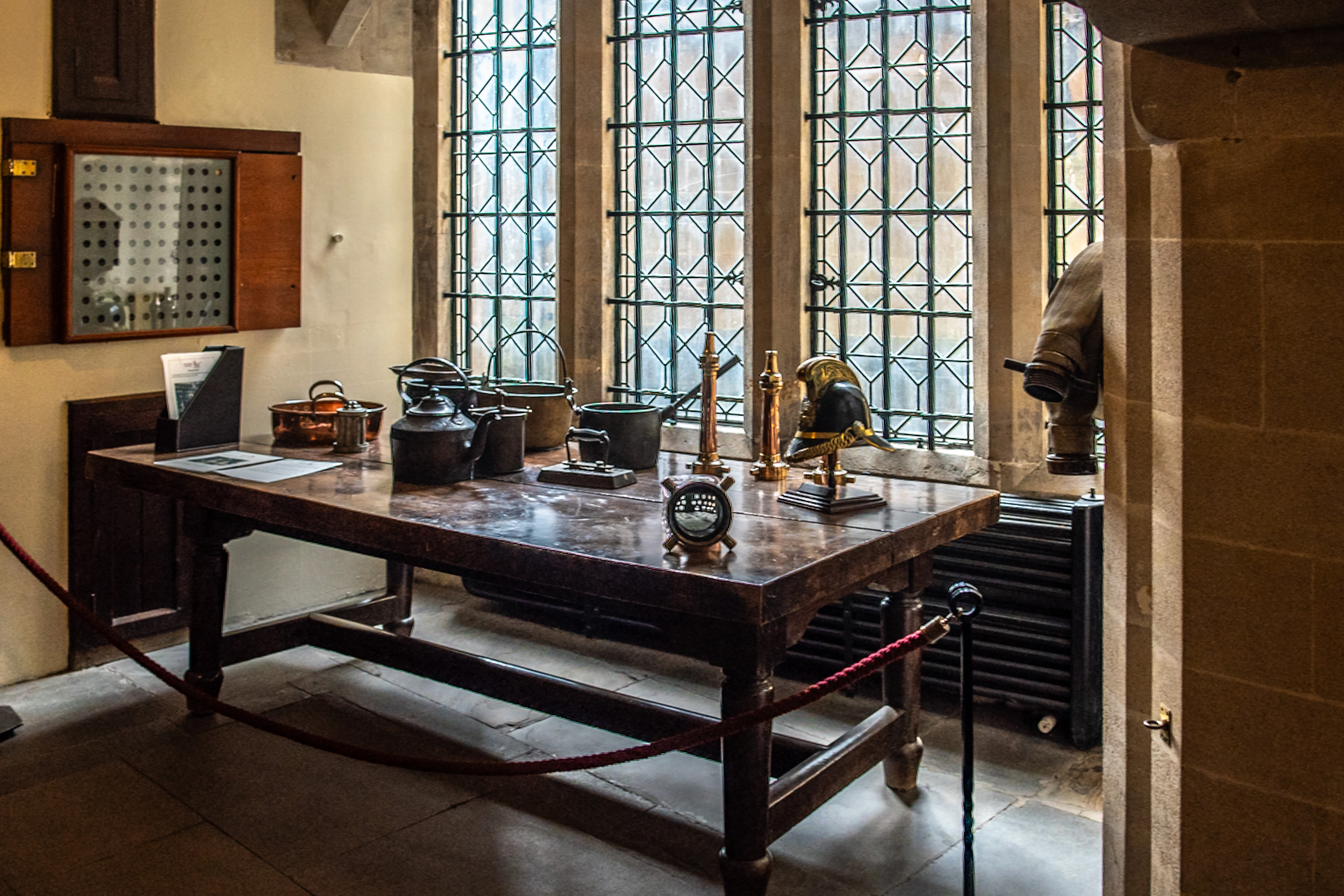 A wooden table is laden with various antique kitchen items including teapots, pots, and an iron. Behind the table, a large window with geometric panes lets in natural light, and a wooden cabinet with a perforated door hangs on the wall.