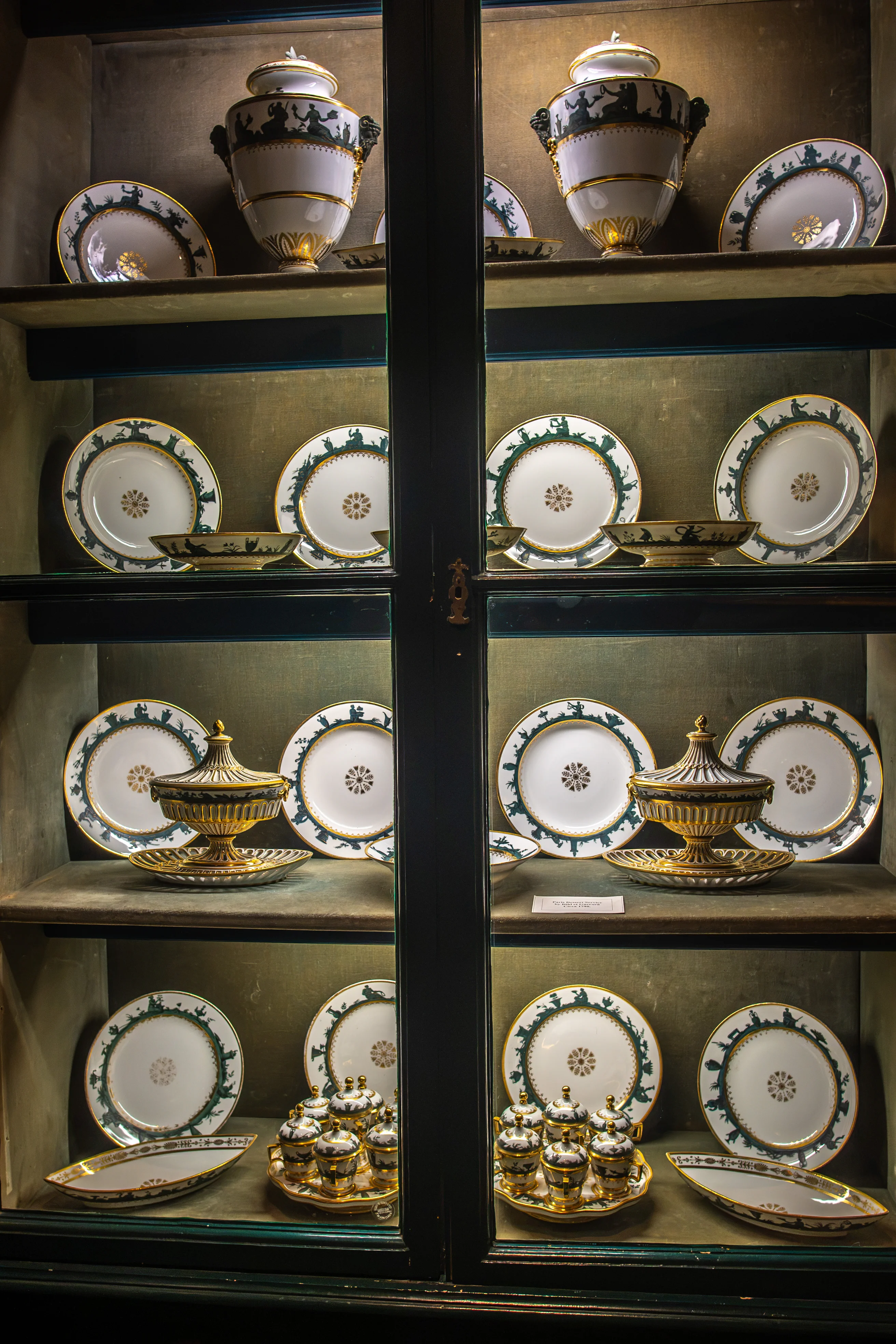 A display cabinet filled with antique white porcelain plates, serving dishes, and a lidded urn decorated with green silhouettes and gold trim.