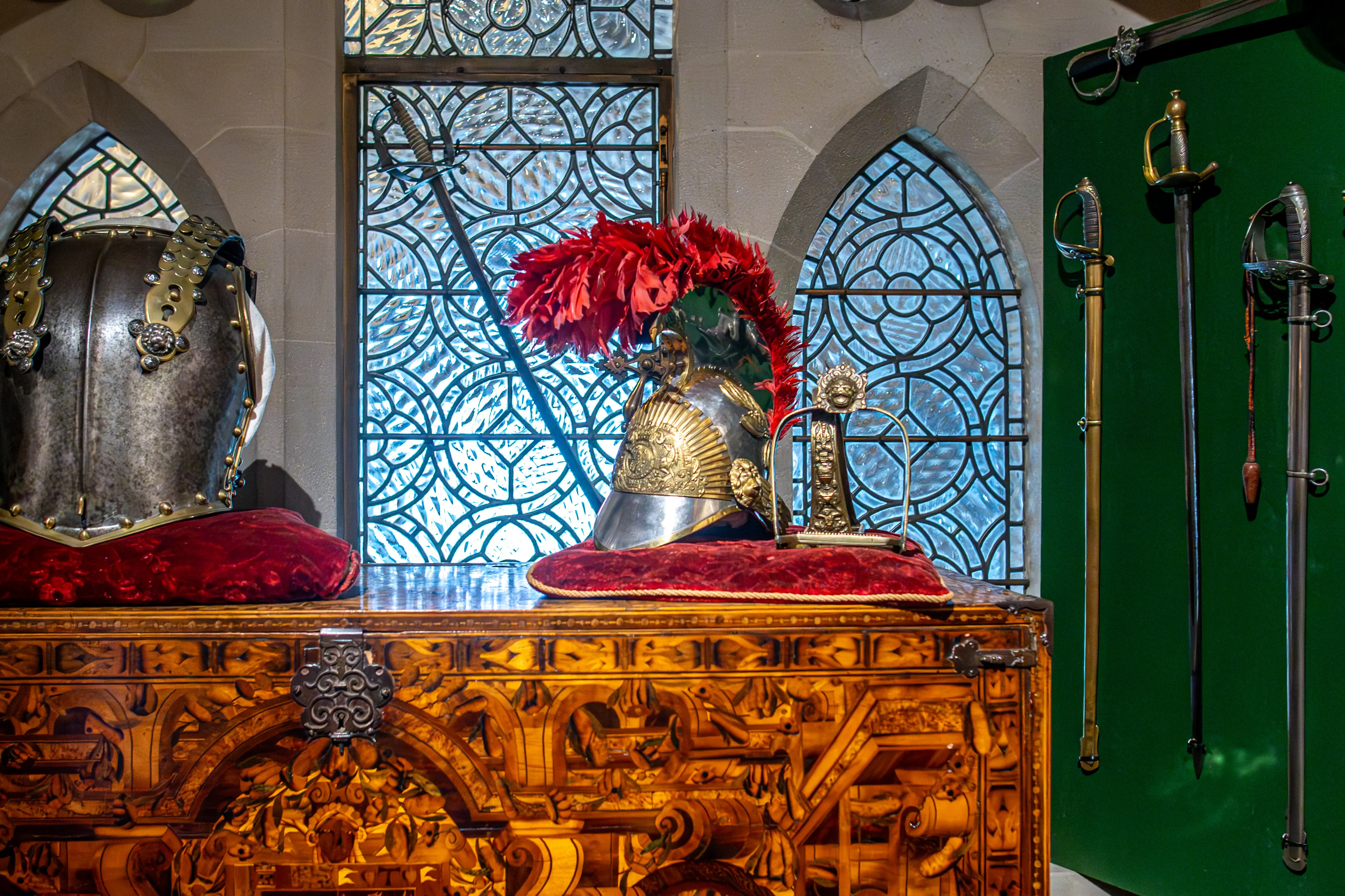A close-up view of an ornate, intricately carved wooden chest, topped with two ceremonial helmets on red cushions, with a collection of swords mounted on a green wall in the background.