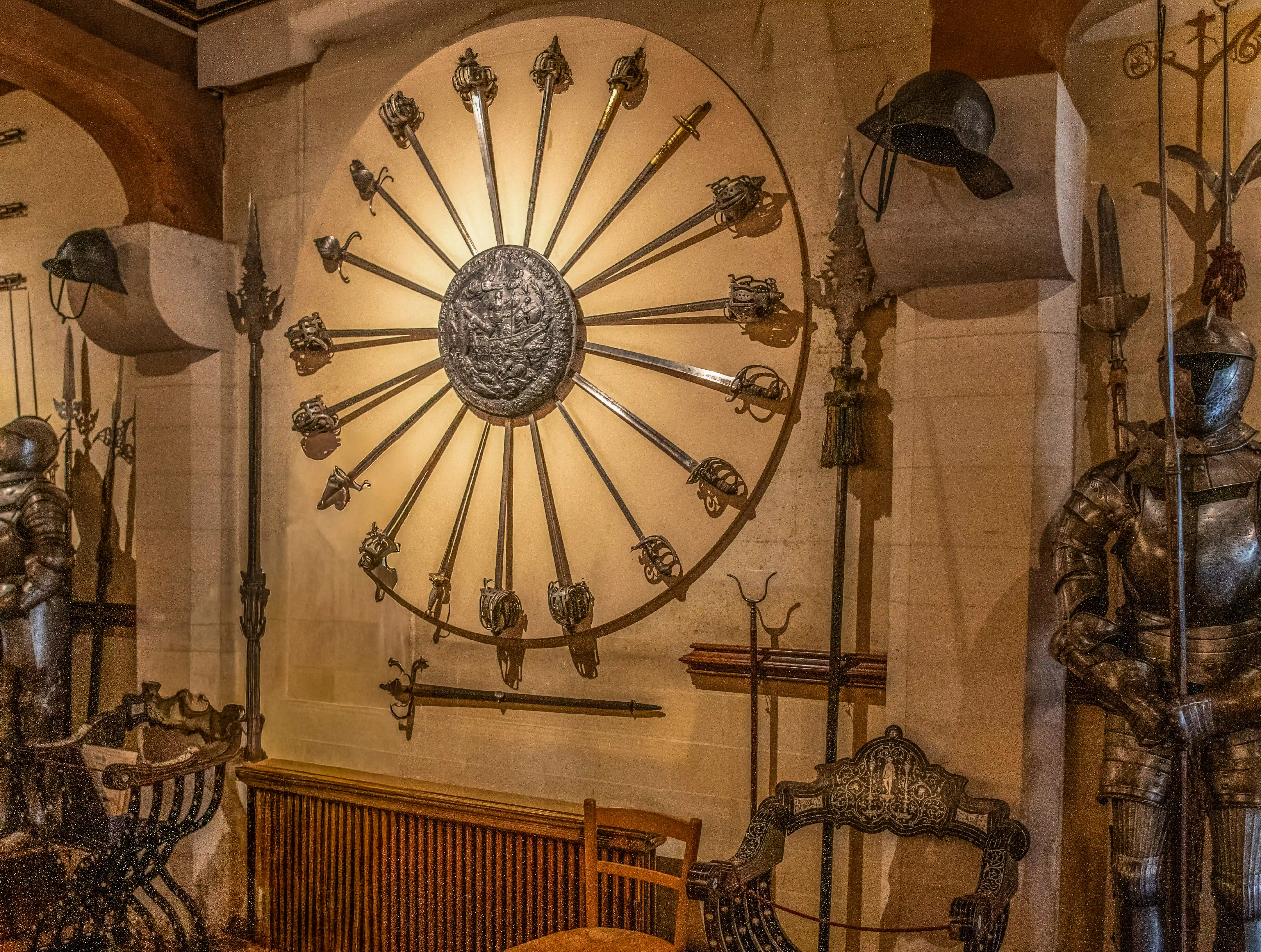 A room filled with antique weapons and armour. A large circular display on the wall features numerous swords radiating from a central medallion. To the right, a full suit of armour stands guard, and to the left, another suit is partially visible. Various polearms and helmets are also on display.