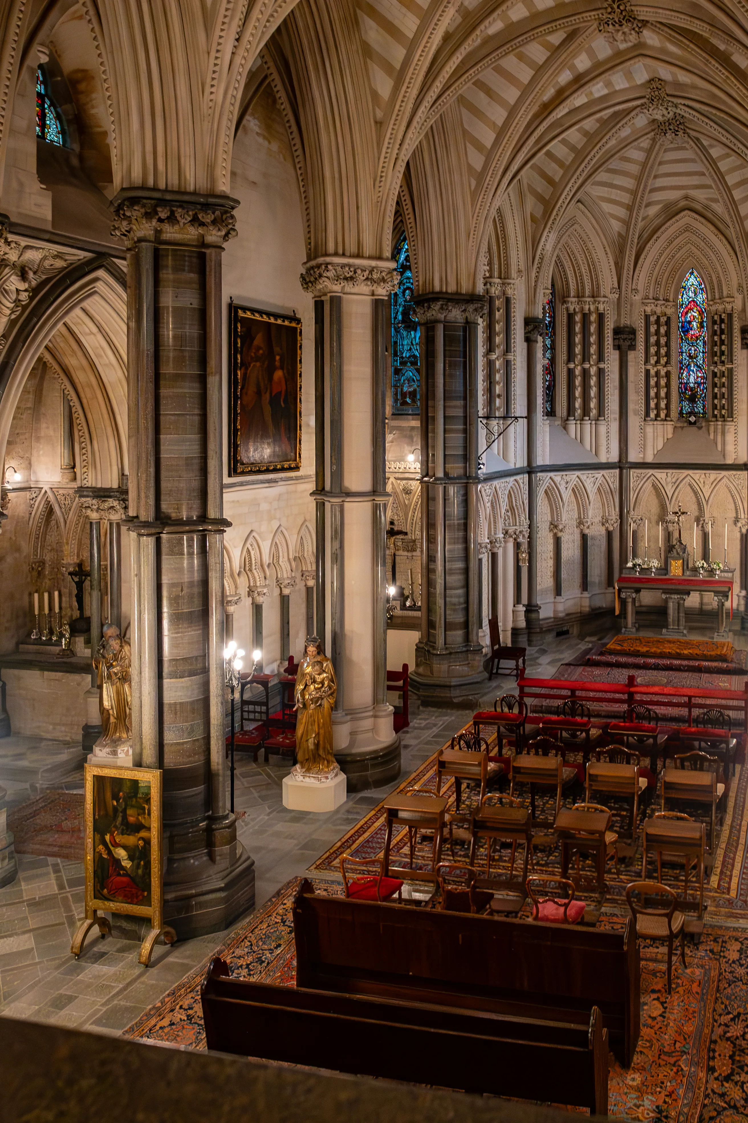 Interior of a Gothic cathedral, looking towards the altar, featuring high vaulted ceilings, stone columns, stained-glass windows, pews, and ornate carpets.