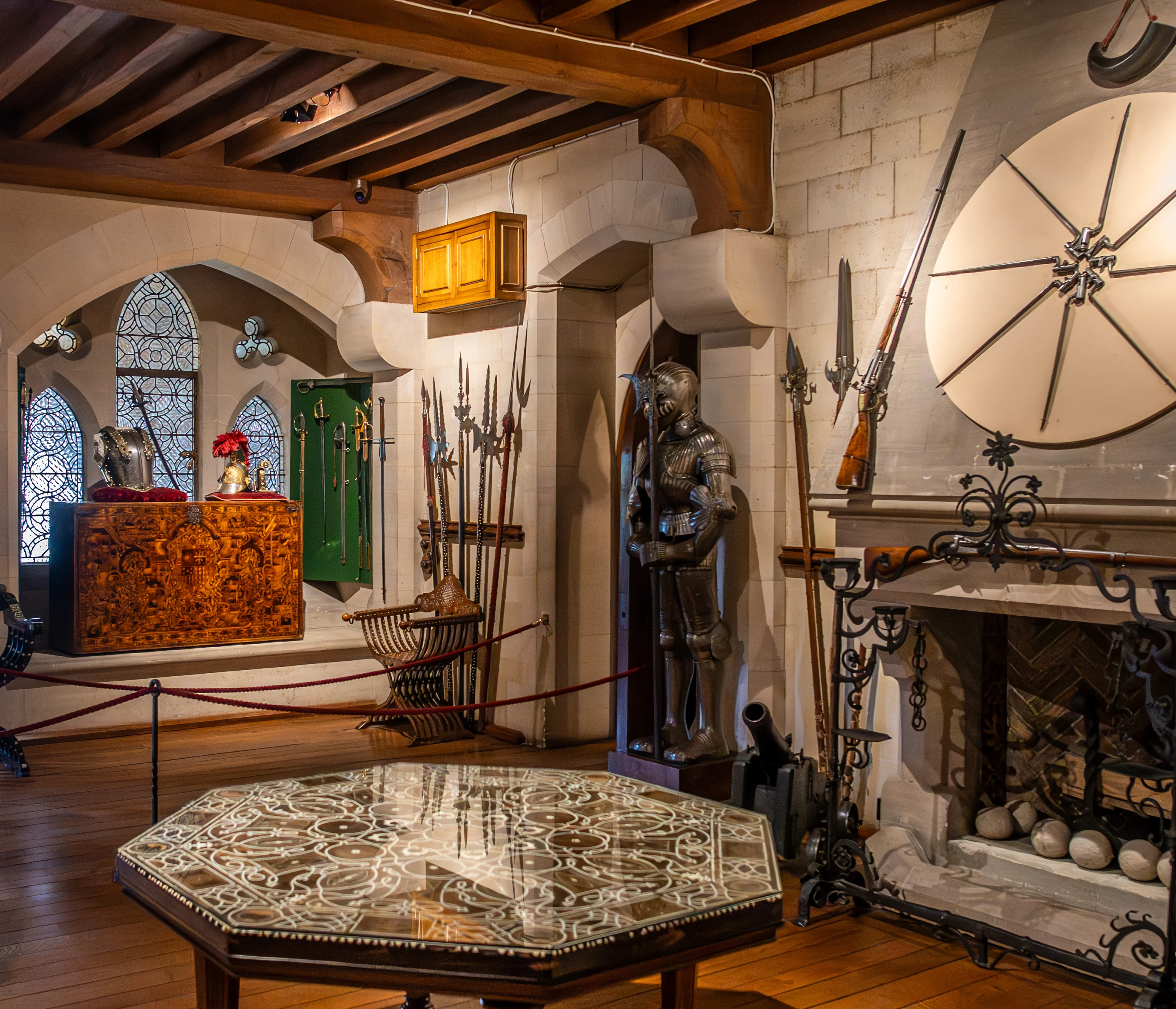 Interior view of a room displaying historical armour and weapons, featuring a suit of armour, spears, rifles, a cannon, a large ornate chest, and an inlaid octagonal table.
