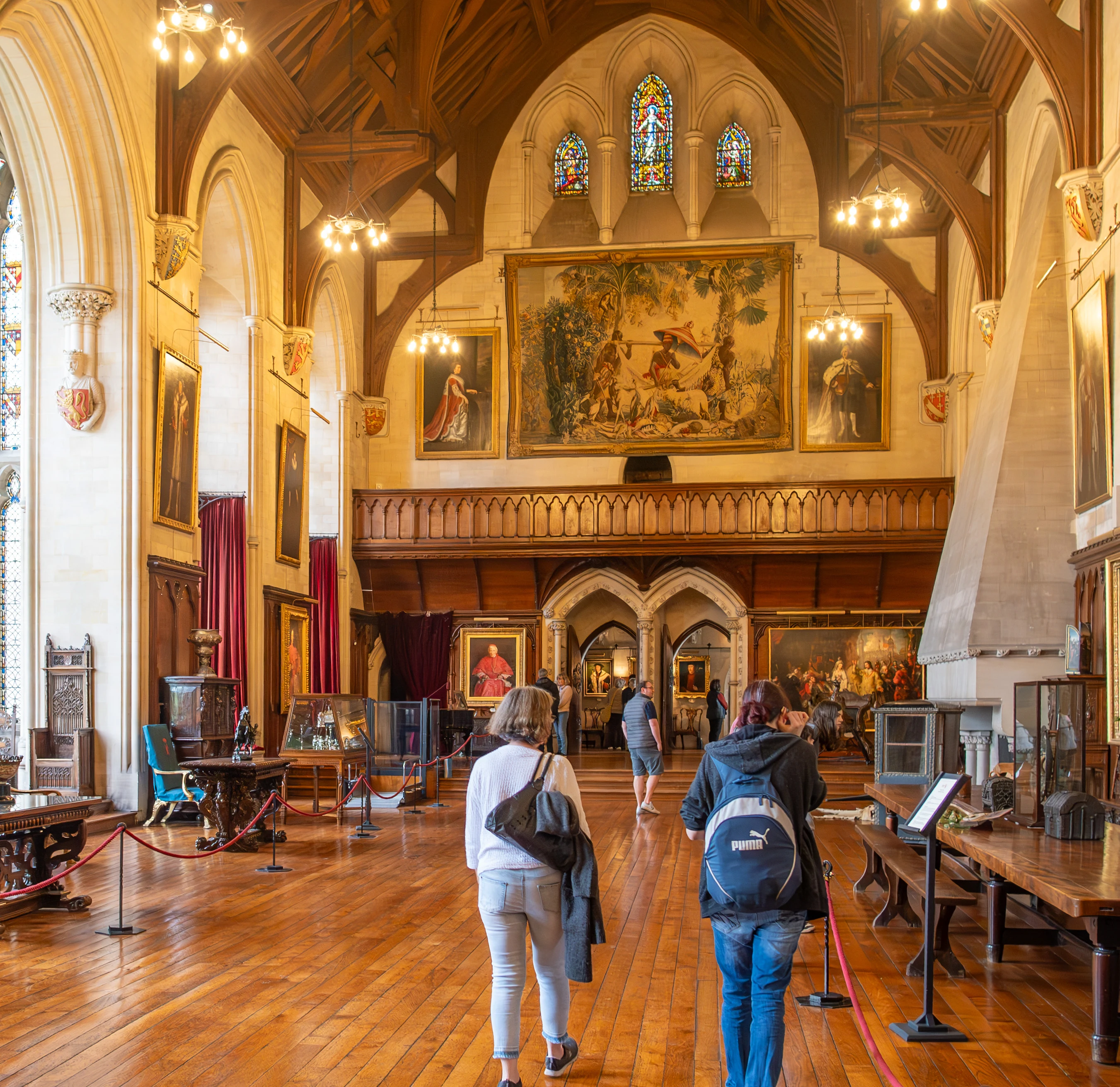 Two people walking away from the camera through a large, ornately decorated hall with high ceilings, wooden beams, stained glass windows, several large framed paintings, and a large tapestry on the far wall.