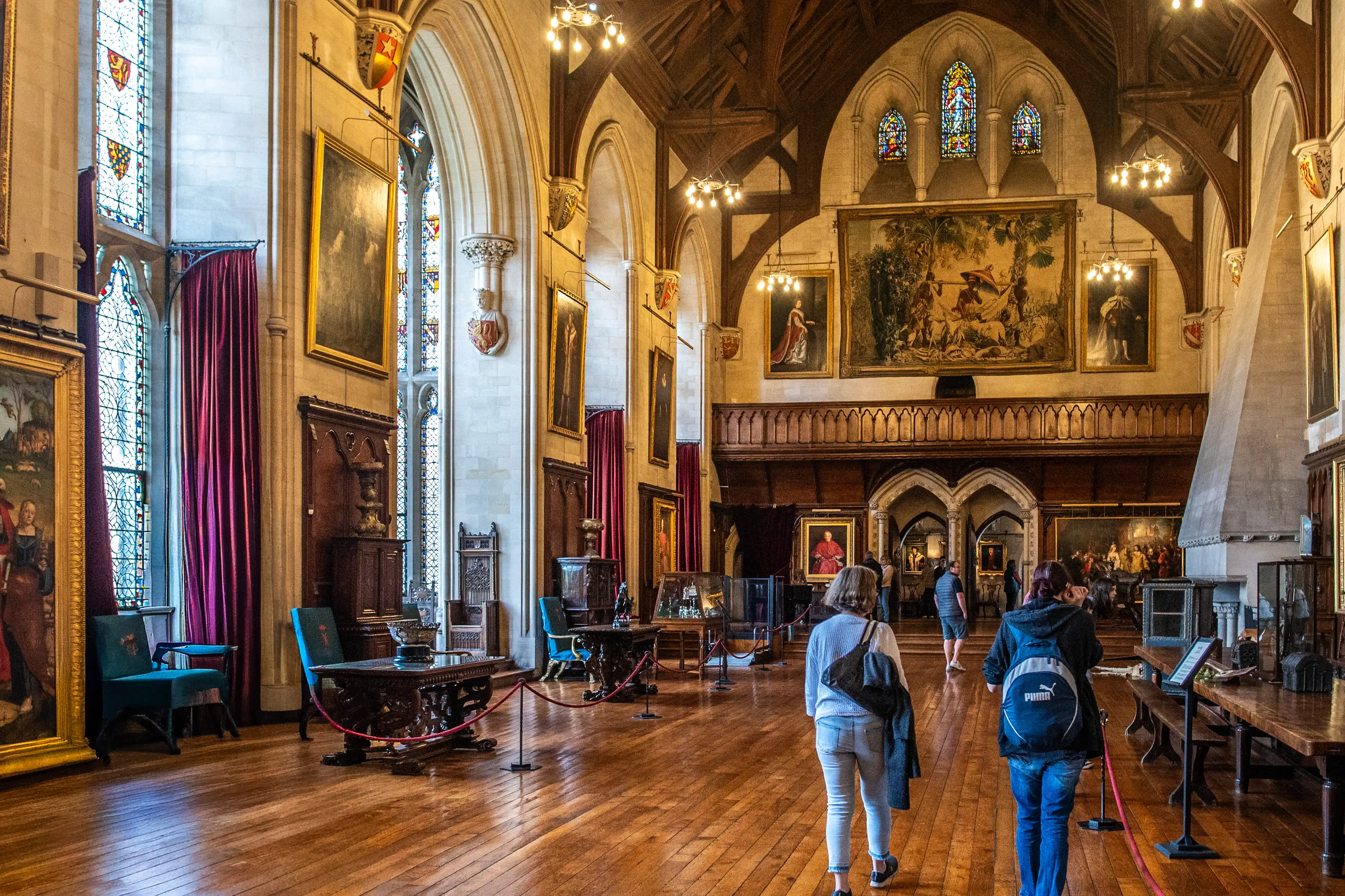 Two people walking away from the camera through a large, ornately decorated hall with high ceilings, wooden beams, stained glass windows, several large framed paintings, and a large tapestry on the far wall.