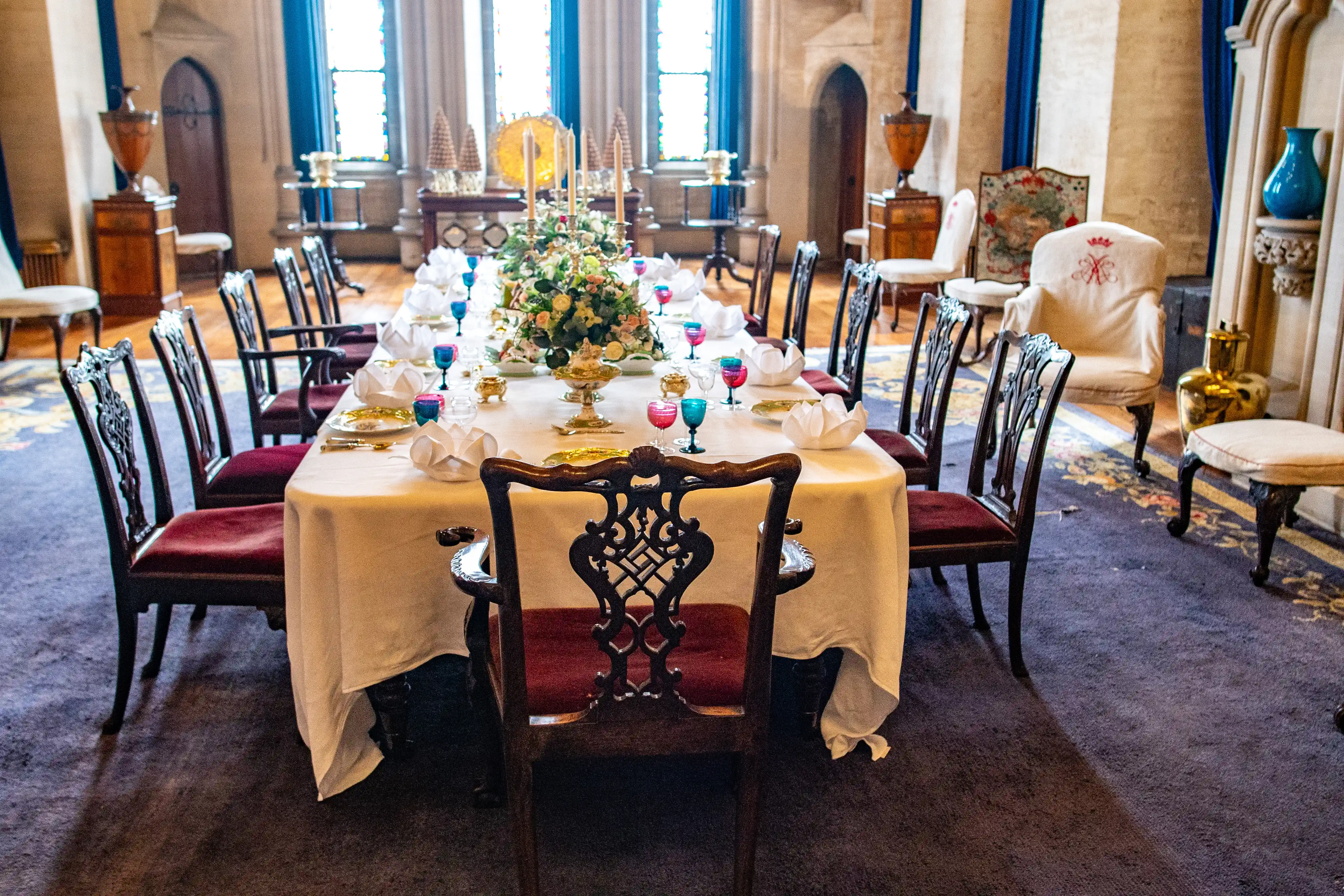 A long dining table draped in a white tablecloth, laden with place settings for a formal meal. Multiple dark wood chairs with red velvet upholstery surround the table, which is adorned with a large floral arrangement and colourful glass goblets. In the background, large windows and antique furnishings create a sense of opulence.