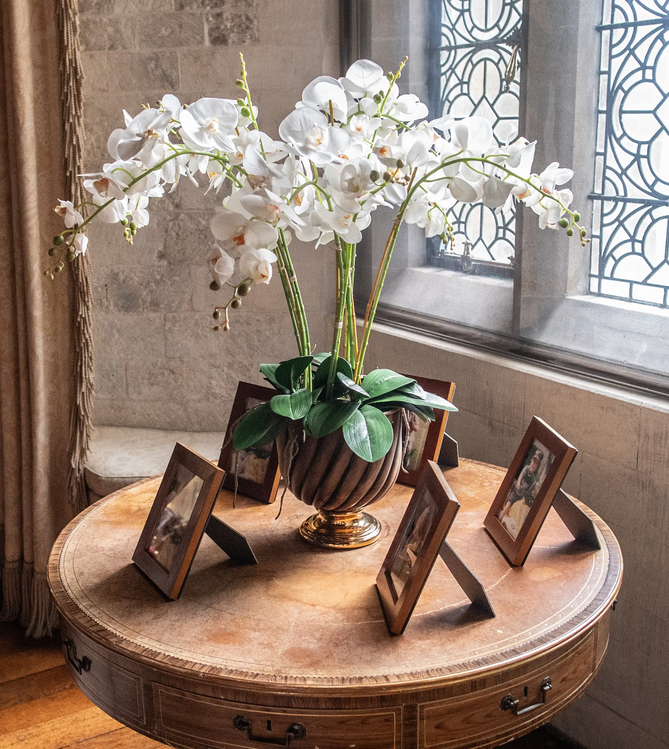 A stunning arrangement of white orchids in a metallic fluted vase, placed on a round vintage wooden table. The table also holds two framed photographs. The backdrop features a stone wall and a window with intricate latticework