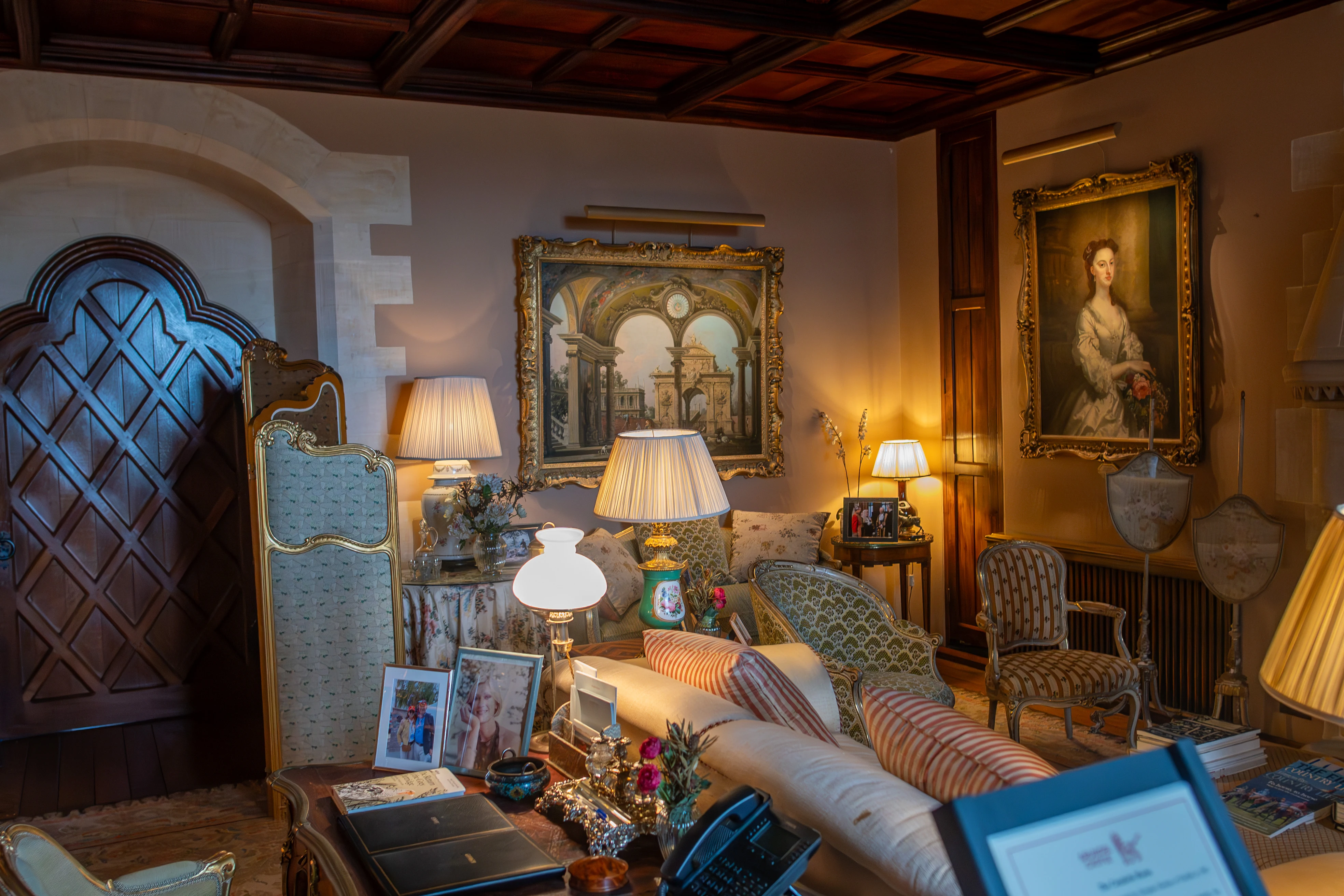 An interior view of a traditional drawing room with antique furniture, including a sofa and chairs, adorned with patterned upholstery and cushions. Several framed paintings, lamps, and decorative items are displayed around the room, illuminated by warm lighting. A large wooden door with a diamond pattern is visible on the left.