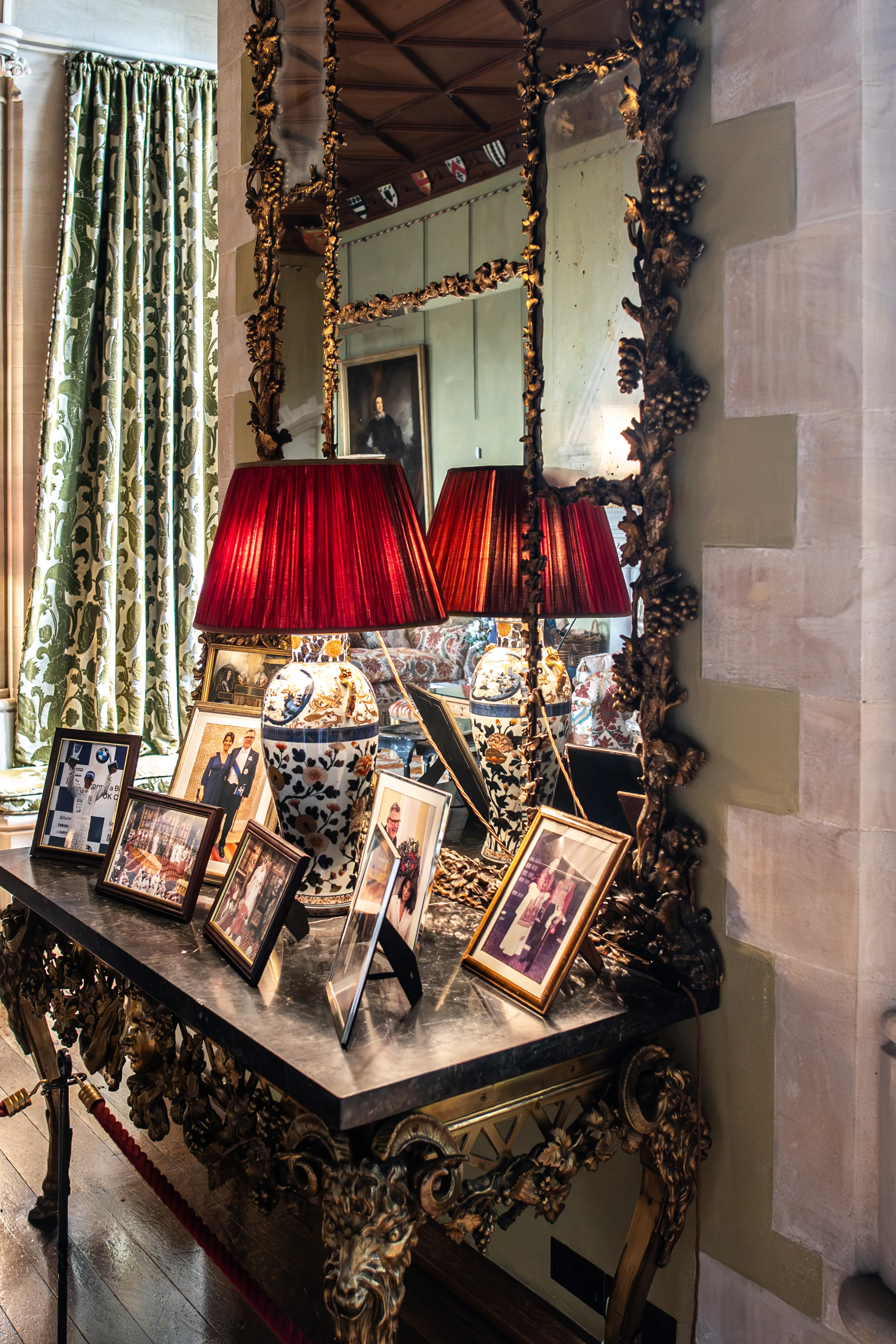 A close-up of a dark marble-topped console table in an ornate room. The table holds several framed photographs and two red-shaded lamps with decorative ceramic bases. A large, gilded mirror with a vine motif reflects the room, including a portrait and patterned curtains.