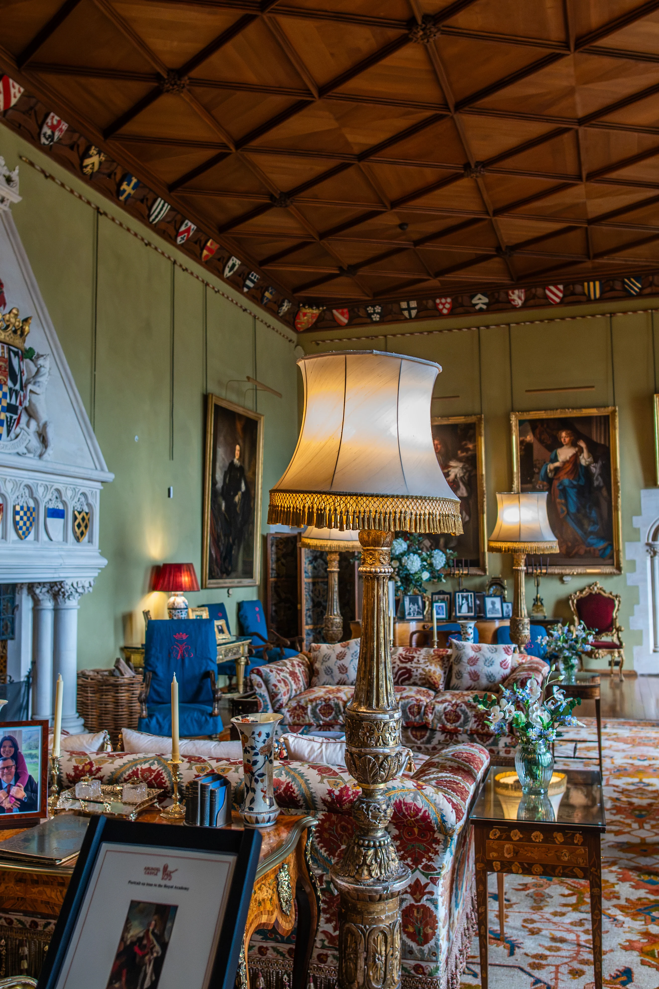 An interior view of a lavish drawing room, showcasing a large white fireplace with escutcheons, patterned sofas and armchairs, ornate floor lamps, and classical portraits hanging on pale green walls.