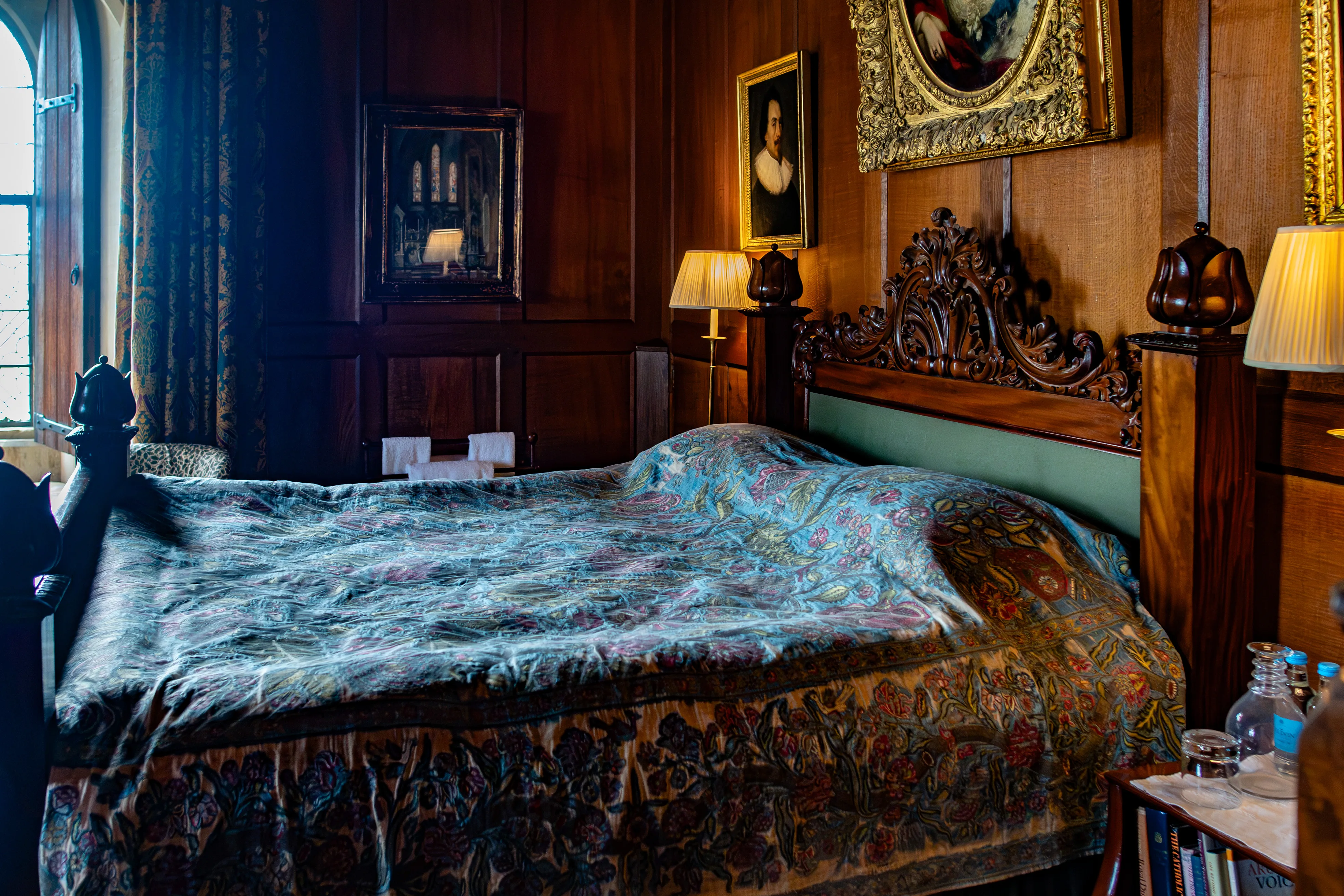 A vintage bedroom with a large wooden bed featuring an ornate headboard and a patterned bedspread in blue, green, and gold. The room has wooden paneled walls, framed artwork, antique lamps with pleated shades, and a window with dark curtains. A bedside table holds a glass of water and a book.