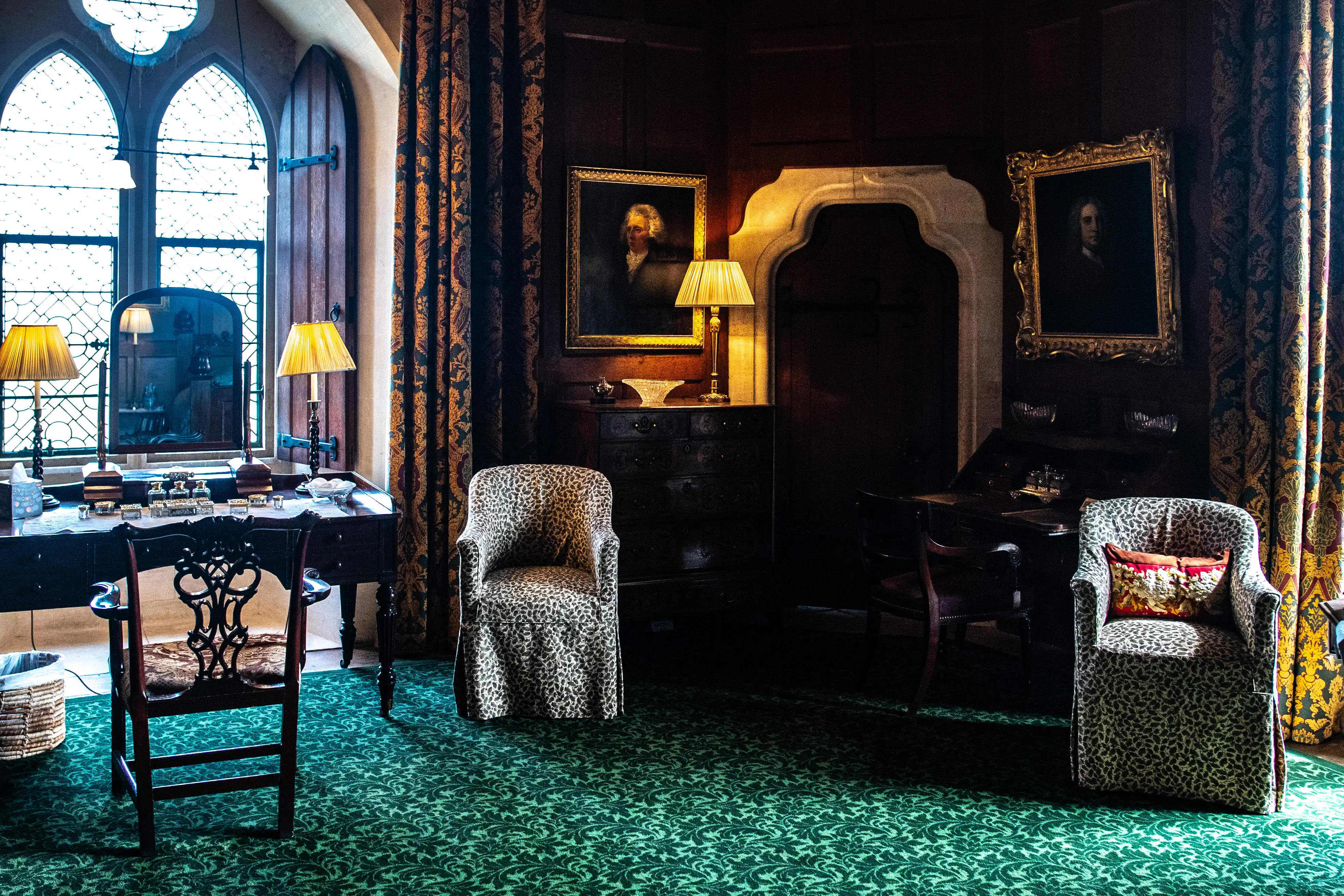 A luxurious, dimly lit drawing room with a prominent green patterned carpet. Two armchairs with leopard print upholstery are placed centrally. A writing desk with a mirror and decorative items is positioned near a large gothic window. Two framed portraits hang on the dark wood-panelled walls, and ornate curtains are visible.
