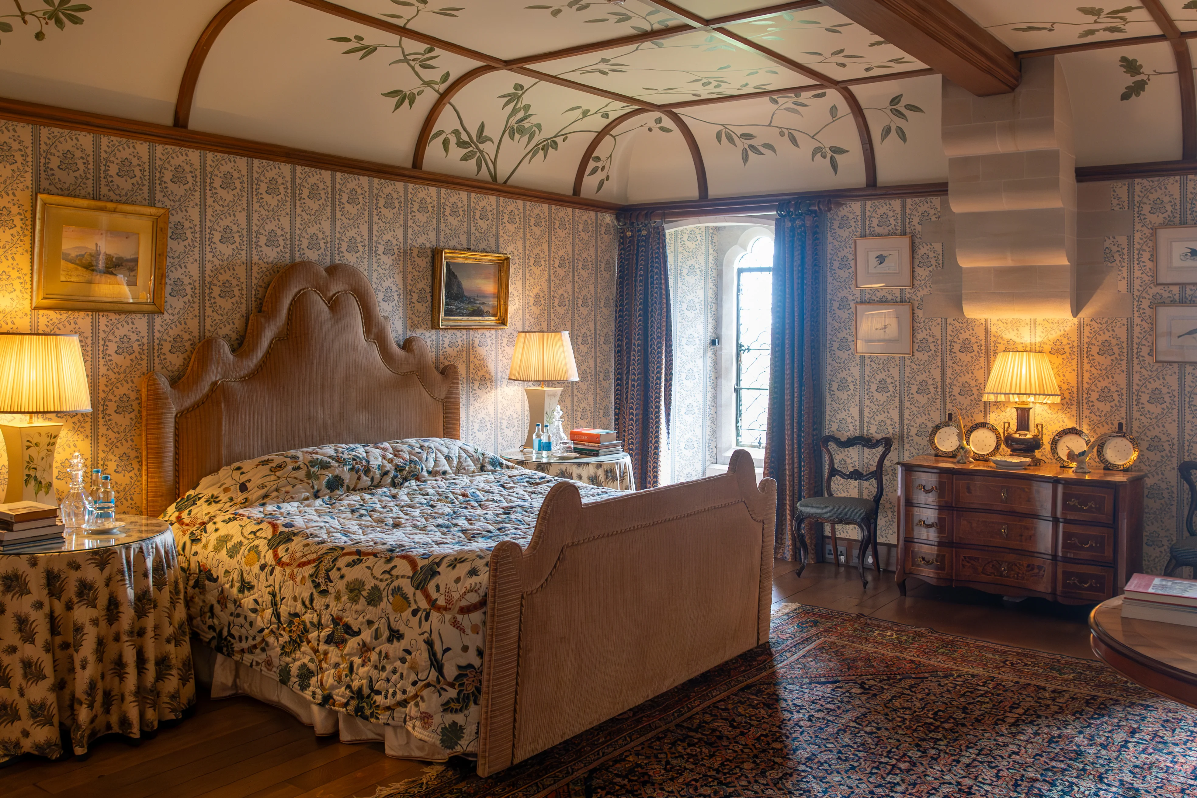 A luxurious bedroom interior with a large bed covered in a floral quilt, decorative wallpaper, a vaulted ceiling painted with greenery, and antique furniture.