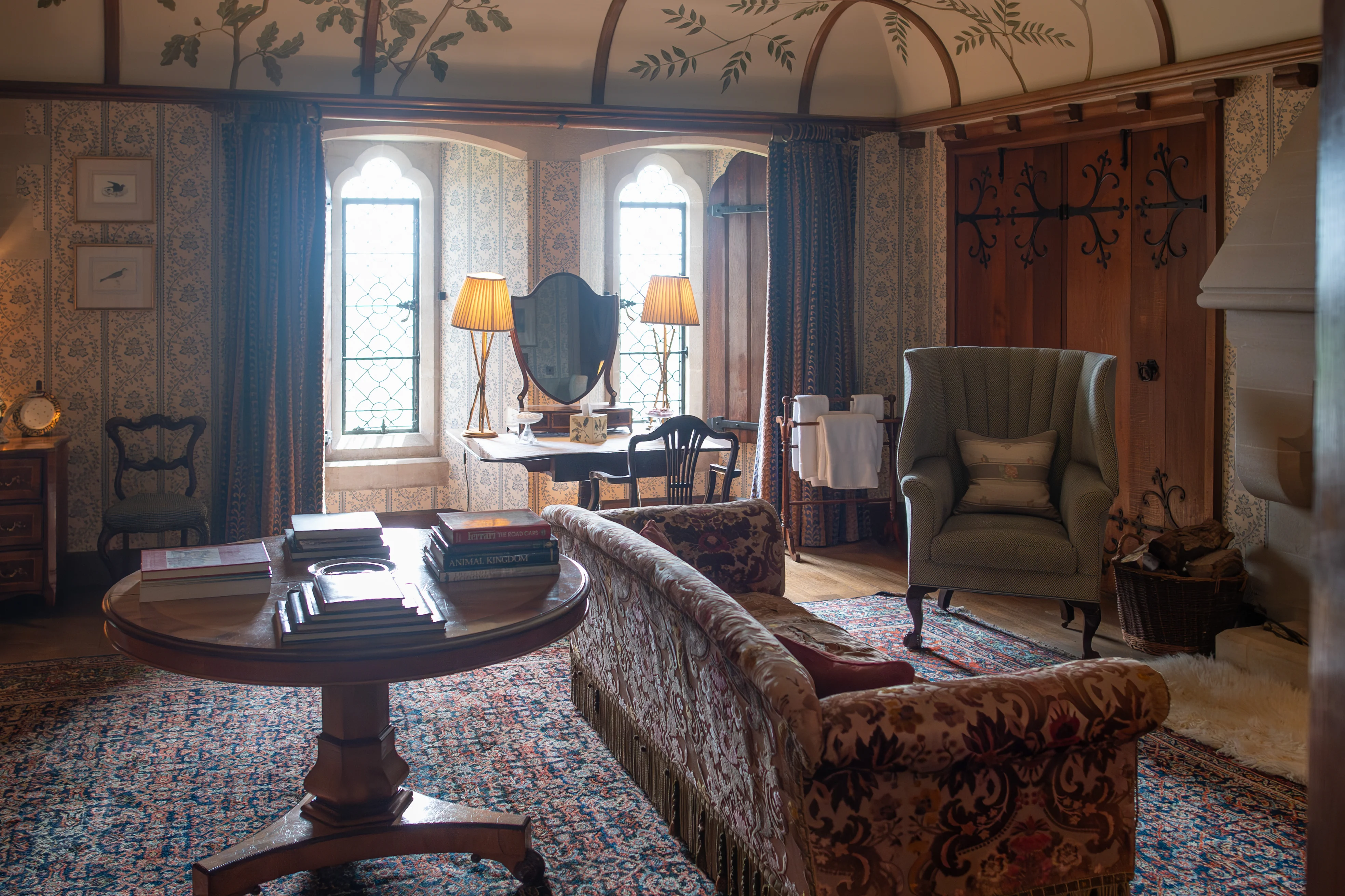 Interior of a luxurious drawing room with a patterned sofa, a green wingback armchair, a dressing table with two lamps and a mirror, a round wooden table with books, a patterned rug, and windows with gothic arches. The ceiling is vaulted and decorated with painted branches and leaves.