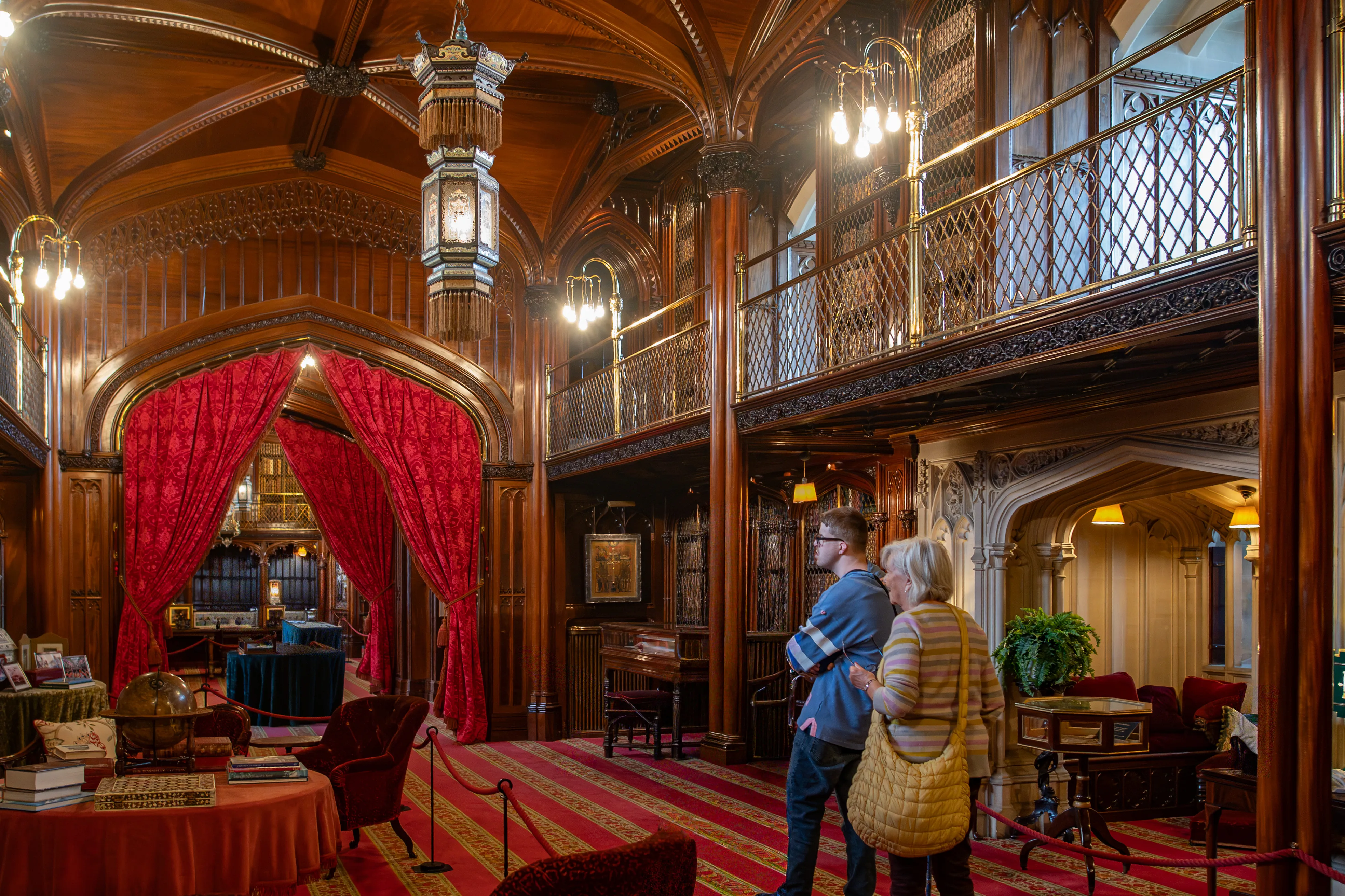 Three people stand in a large, richly decorated library, looking towards an arched doorway draped with red velvet curtains. The library has dark wood panelling, a mezzanine level with bookshelves, and elaborate chandeliers