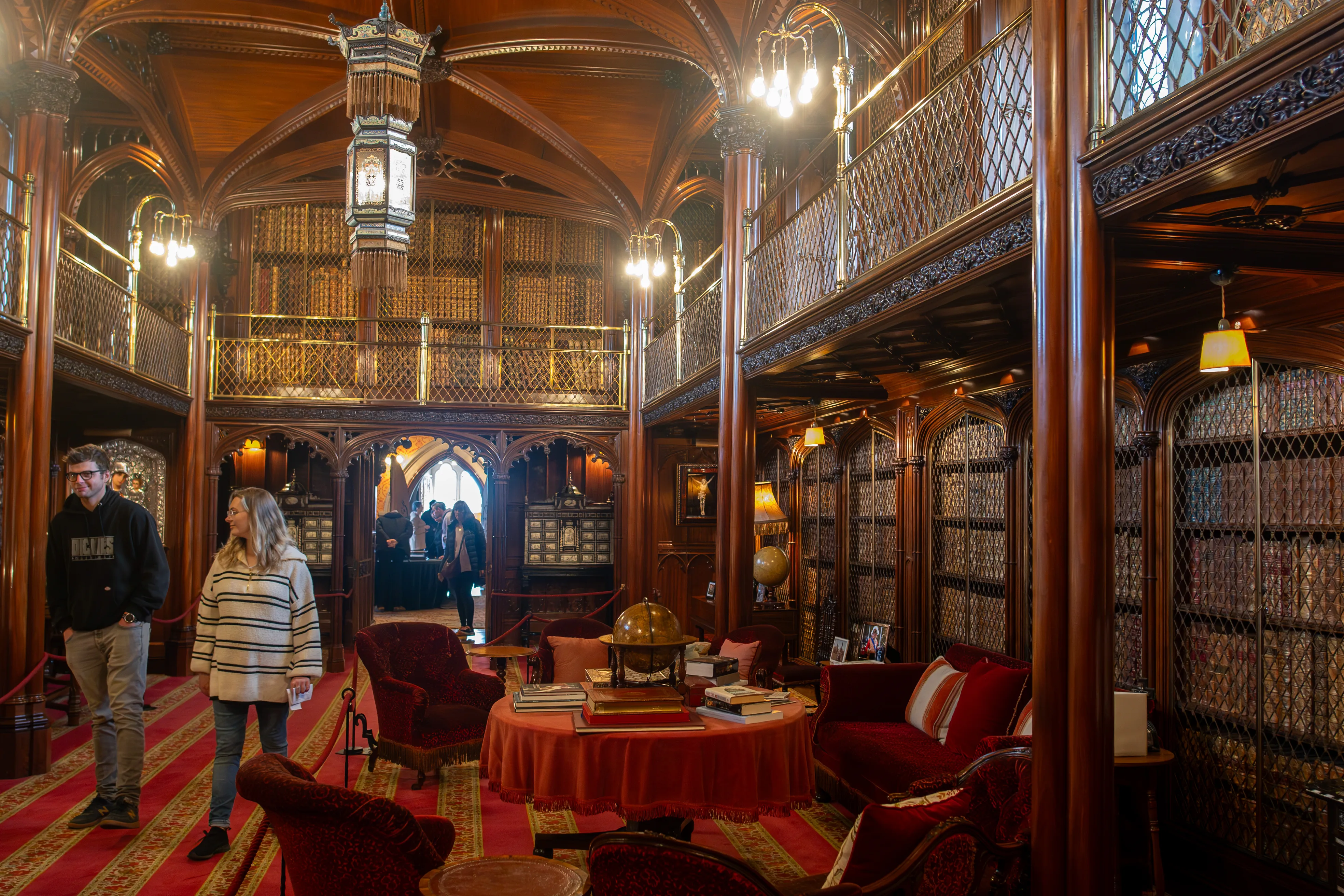 A wide shot of an ornate library with dark wooden shelves reaching to the ceiling, featuring multiple levels and intricate detailing. Red velvet armchairs and sofas are arranged around a central table, and two people are walking through the room.