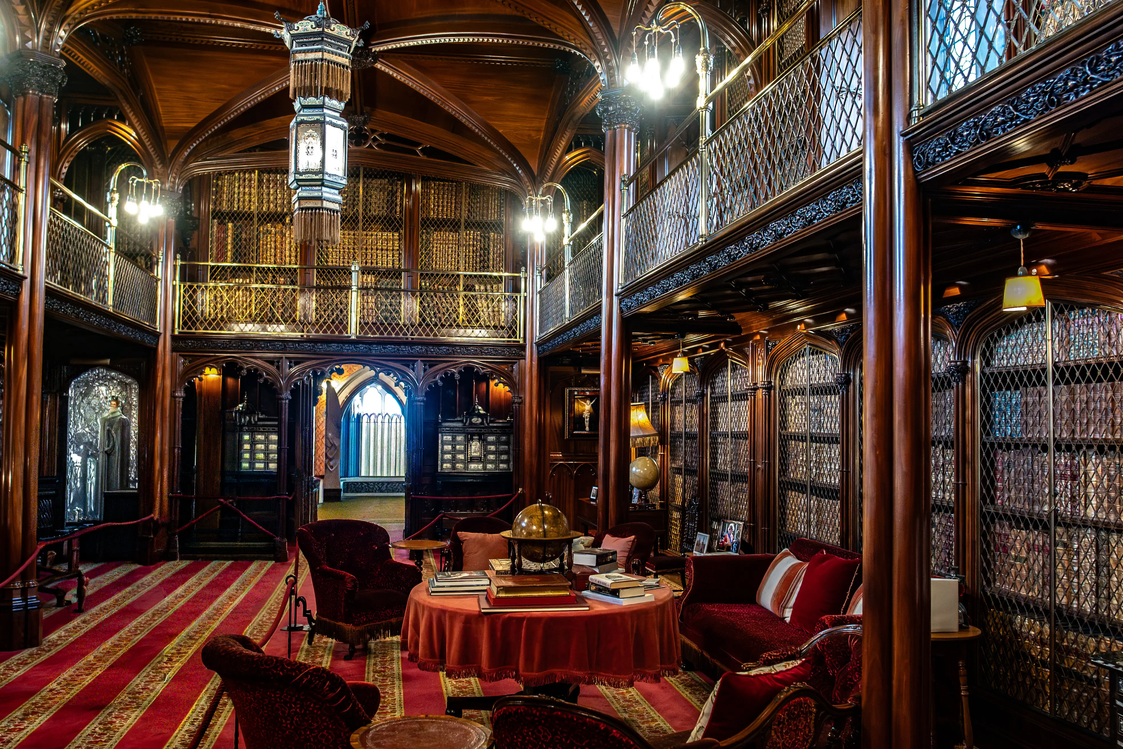 A wide shot of an ornate library with dark wooden shelves reaching to the ceiling, featuring multiple levels and intricate detailing. Red velvet armchairs and sofas are arranged around a central table, and two people are walking through the room.
