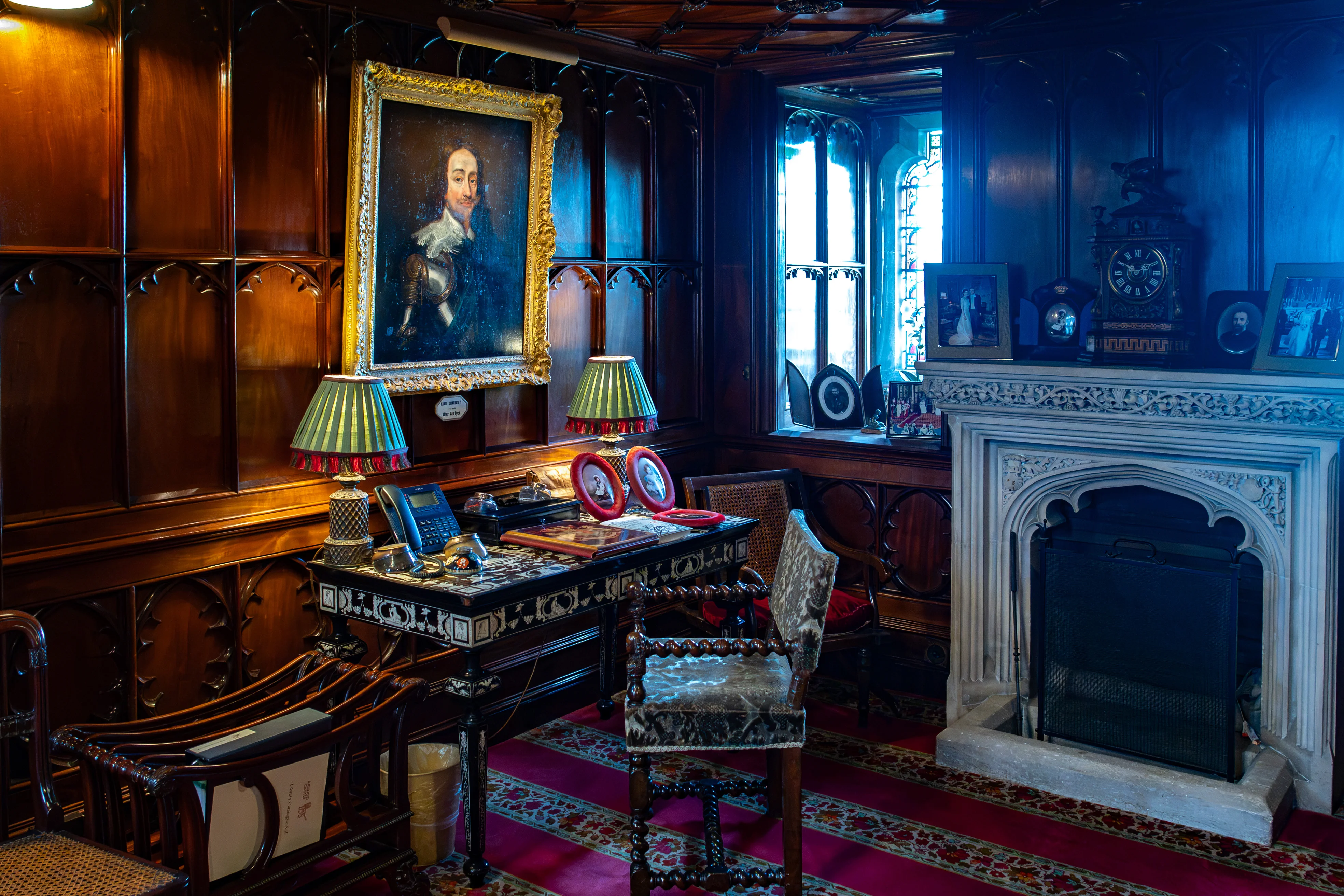 A detailed view of a traditional study room, showcasing a large oil painting of a man in historical attire, a dark wooden desk with a landline telephone and two framed pictures, ornate lamps with fringed shades, a decorative fireplace with a carved mantelpiece and a large clock, and a brightly coloured rug.