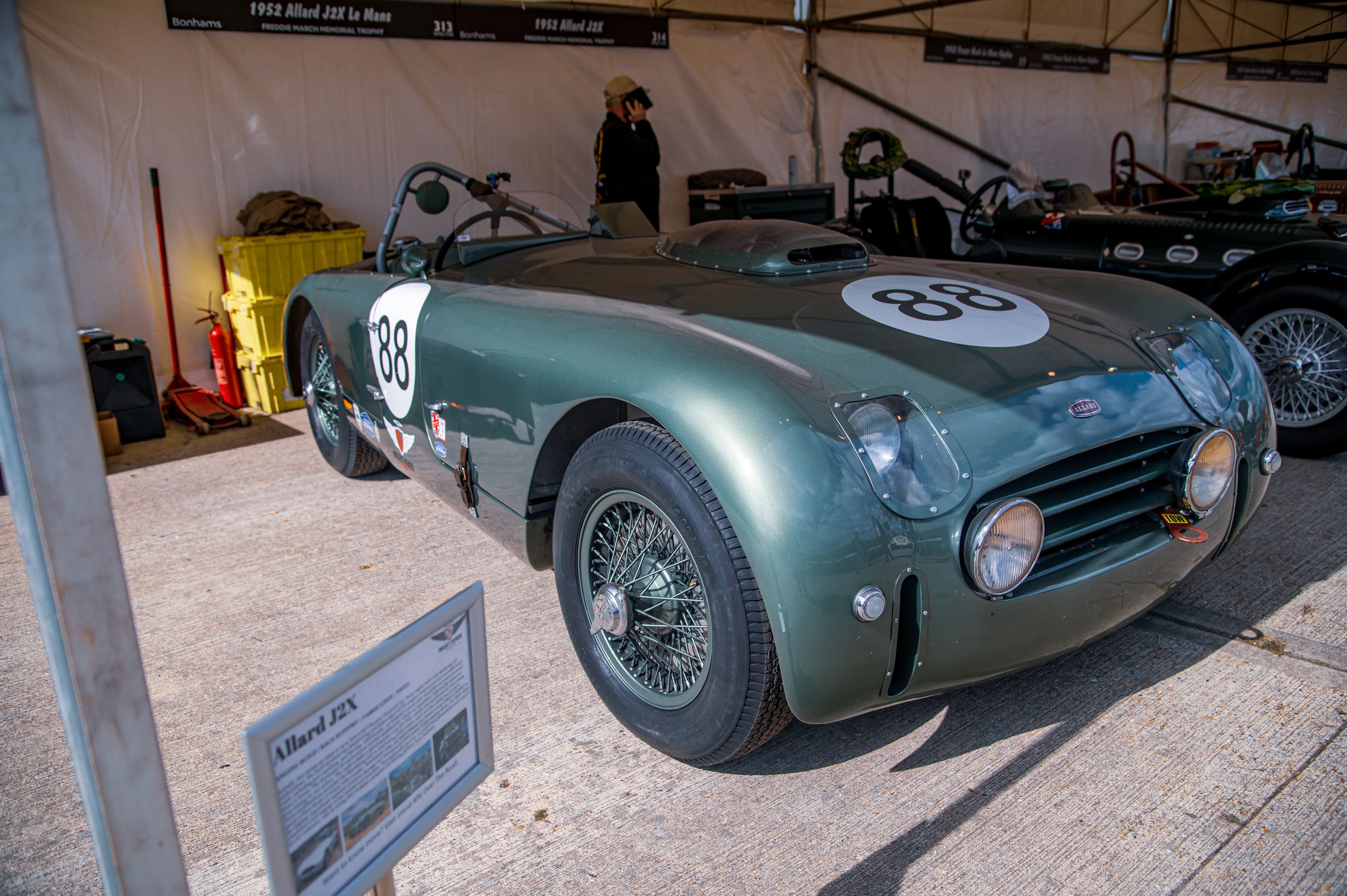 A close-up of a vintage green Allard J2X race car with the number 88 prominently displayed on its bonnet and side. The car features spoked wheels and a low-slung profile. A small informational sign is visible in the foreground.