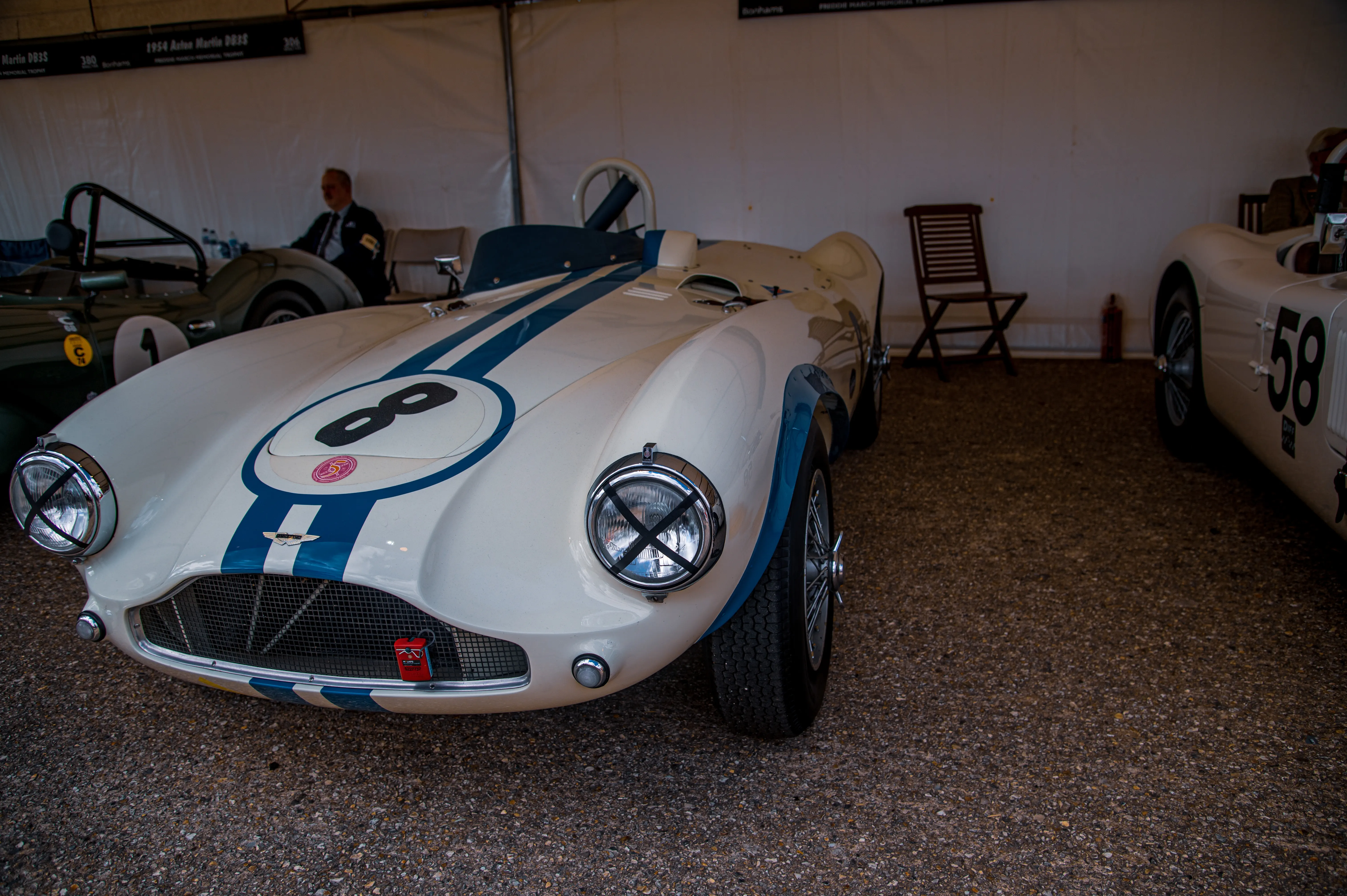 A close-up view of the front of a vintage white Aston Martin DBR3S racing car, featuring blue stripes and the number '8' in a circle on its bonnet.